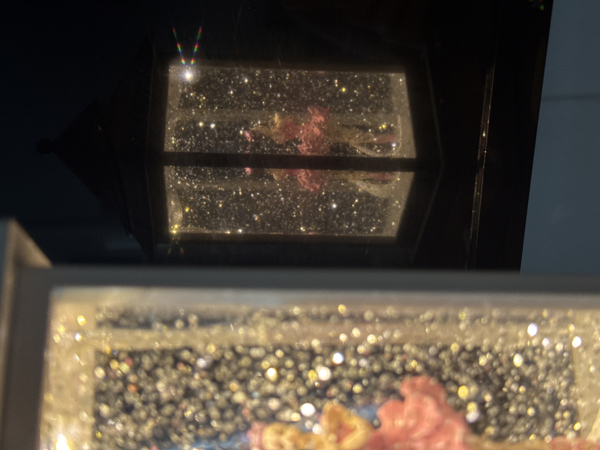 The image shows two blurred snow globes with pink and yellow floral elements inside. The snow globes are filled with glitter. The top snow globe is encased in a dark, lantern-like frame. The background appears to be dark, possibly at night or in a dimly lit room, with a wall partially visible on the right. No information on location could be found.