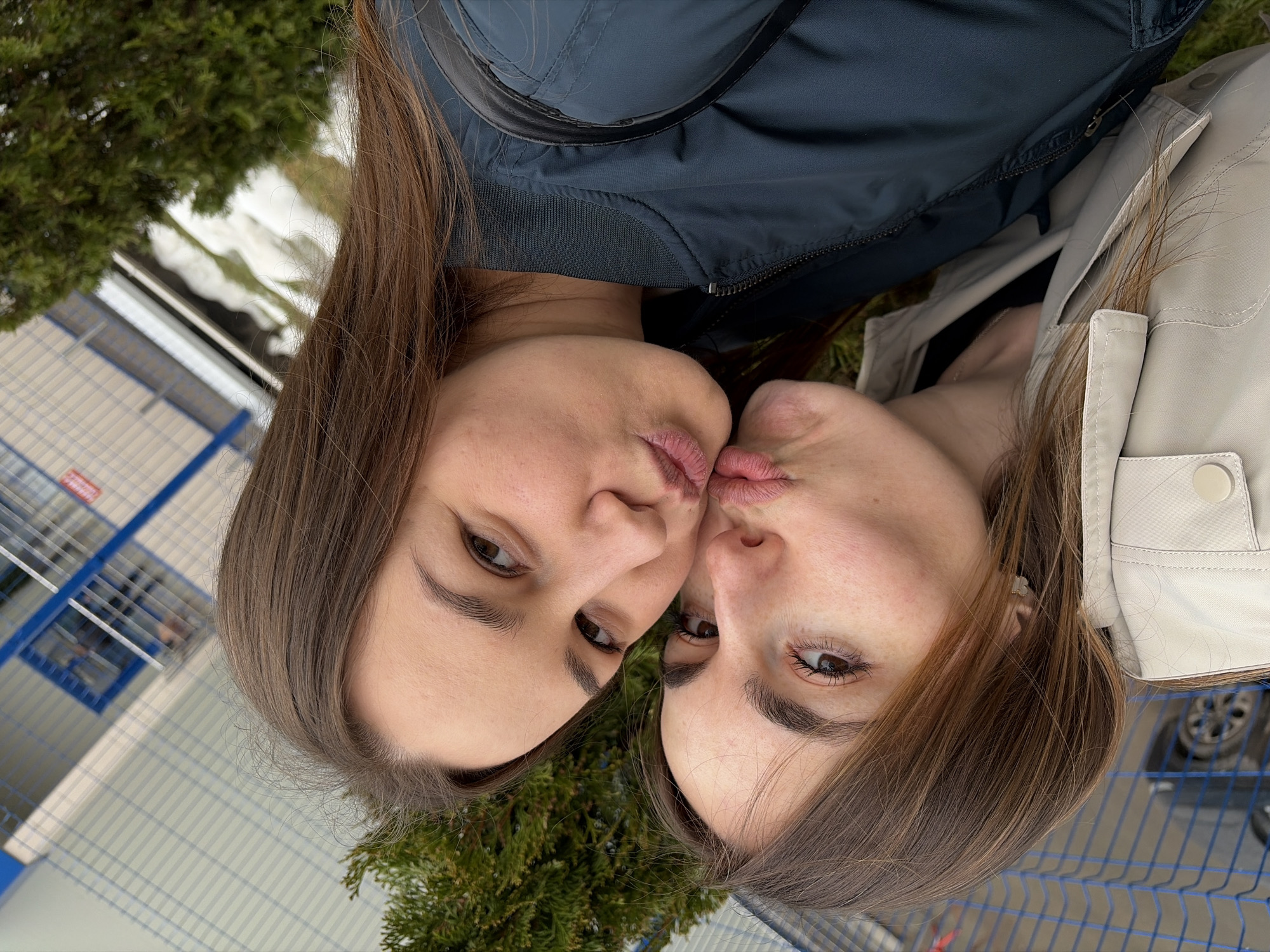 Two young women are positioned face-to-face, almost touching lips, in an outdoor setting. One is wearing a dark blue jacket, while the other has a light-colored jacket. In the background, there is greenery along with structures that resemble a building with blue and white paneling, and a snow patch. Based on the architectural style and the fence, the location might be near a commercial or industrial area. Based on the information given, this image was potentially taken in Izhevsk, Russia.