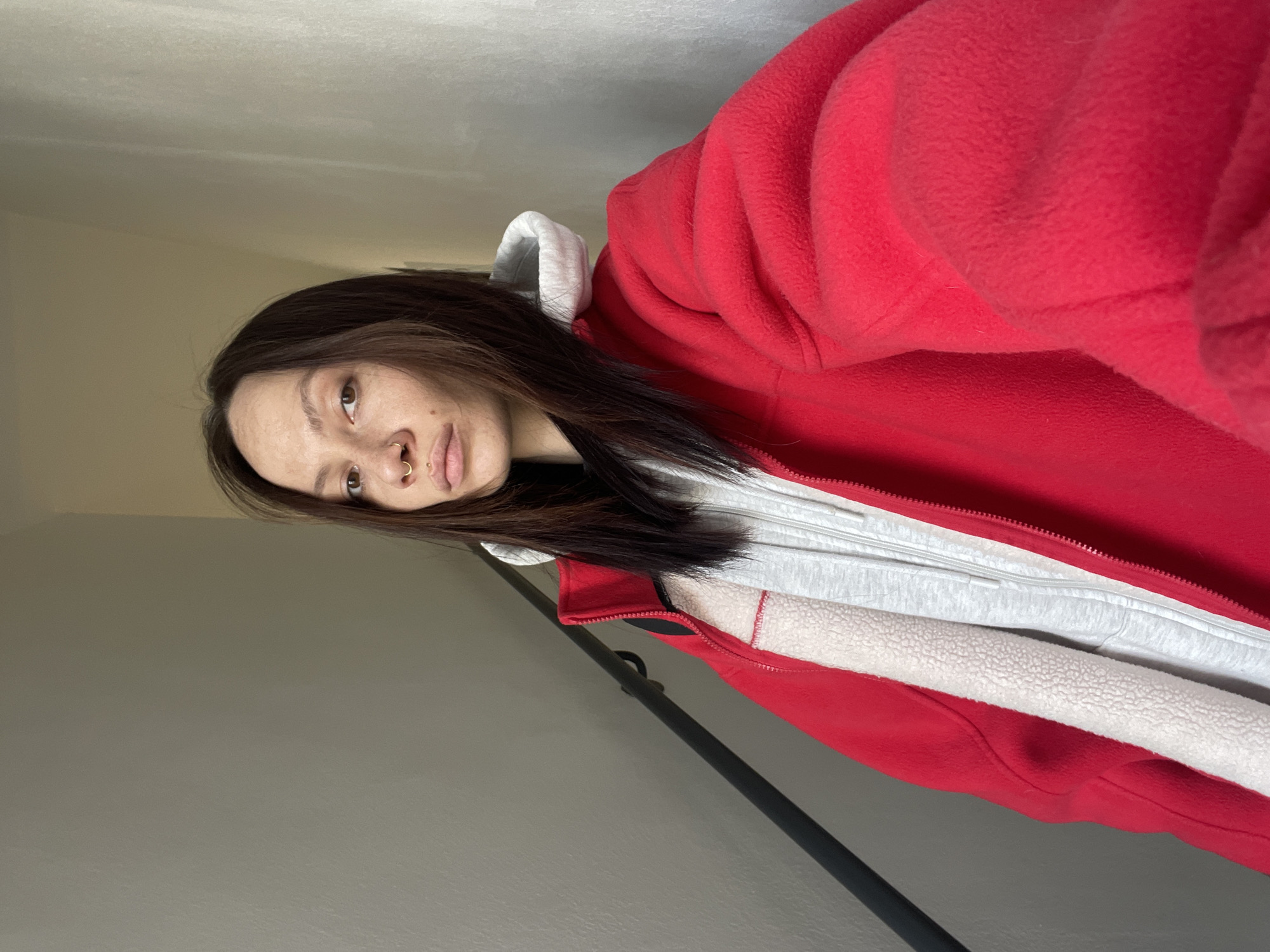 A person, appearing to be female, is depicted in a selfie, with the image rotated 90 degrees counter-clockwise. They have long, dark hair that falls over their shoulders and are looking directly at the camera with a neutral expression. Notable facial features include brown eyes, a septum piercing (gold hoop), a nostril piercing (gold stud), and a labret piercing below the lower lip.
The person is wearing a bright red fleece jacket over a light grey hooded sweatshirt. The red jacket appears thick and soft, possibly an oversized comfort piece, with a visible zipper. The inner lining of the red jacket, partially visible at the opening, is a white, textured sherpa or fleece material. The grey sweatshirt also has a zipper and a visible hood.
The setting appears to be indoors. A plain, light-colored ceiling is visible in the upper portion of the frame, and a solid, light grey or beige wall occupies the background in the lower portion. A dark, thin vertical pole or rod is partially visible behind the person's right arm and shoulder. The overall lighting suggests indoor conditions, possibly from a window or ambient room light. There is no visible text in the image. The warm attire suggests cooler weather, consistent with the location context of Izhevsk, Russia.