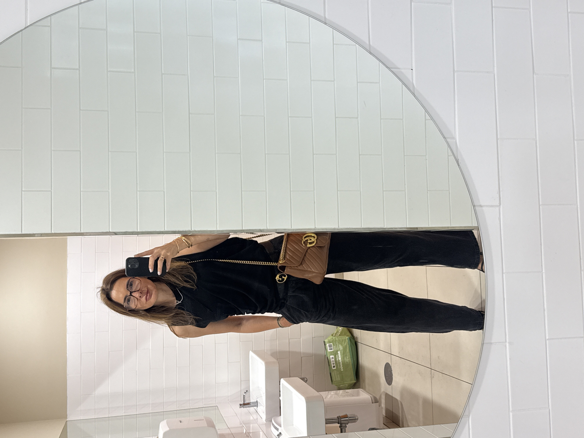 A woman takes a mirror selfie in a public restroom in Vila Nova de Gaia, Portugal. The image is rotated 90 degrees clockwise. She is reflected from the waist up, dressed in a black short-sleeved top and black pants. Her brown hair falls around her shoulders, and she wears dark-framed glasses. She holds a black smartphone in her left hand to capture the photo. Her right wrist is adorned with multiple gold-toned bracelets, and she has rings on her fingers. A brown quilted Gucci Marmont matelassé shoulder bag with a gold chain strap and a prominent "GG" logo is worn across her body, and another "GG" logo is visible on her belt buckle.
The restroom features white subway tiles on the walls, with a section of an adjacent wall painted a light beige color. Below the woman's reflection, two white sinks with silver faucets and dispensers are visible. The floor is covered in light-colored tiles. On the floor to the right of the sinks is a green bag with partially visible text including "COULIAGUAI" and "COMPOSTAGEM" (Portuguese for composting), suggesting it is a compostable waste bag. The woman appears relaxed, displaying a slight smile. The setting indicates a clean, modern commercial establishment.