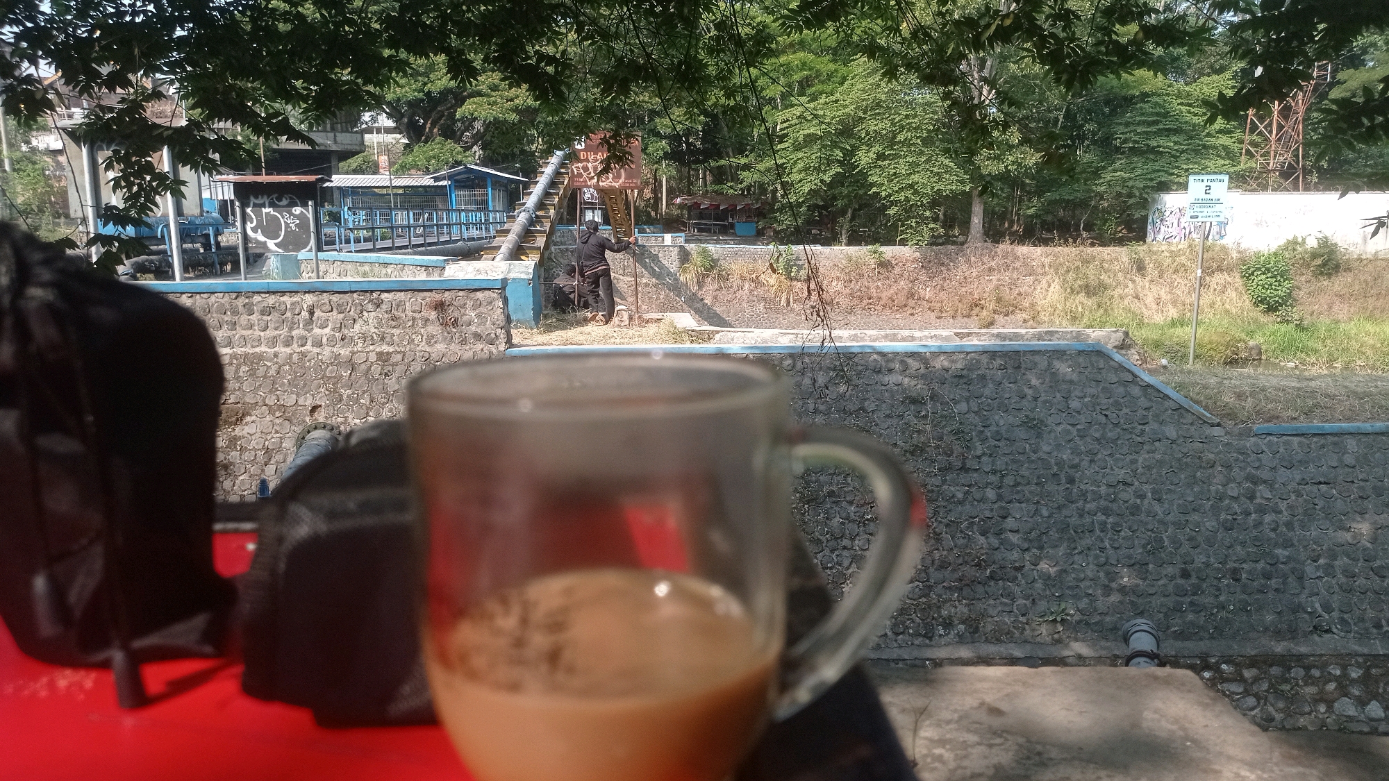 The image shows a close-up view of a glass of coffee on a red surface, with a blurred background of an outdoor environment. The background reveals a stone wall, vegetation, trees, and some structures that appear to be part of a recreational area or park. In the mid-ground, a person in dark clothing is standing near a set of stairs. A sign with text, possibly in Indonesian, is visible in the background, with the number "2" prominently displayed. The scene suggests a casual outdoor setting, likely during the daytime with sunlight casting shadows. The location is identified as Mulyoagung, Indonesia.