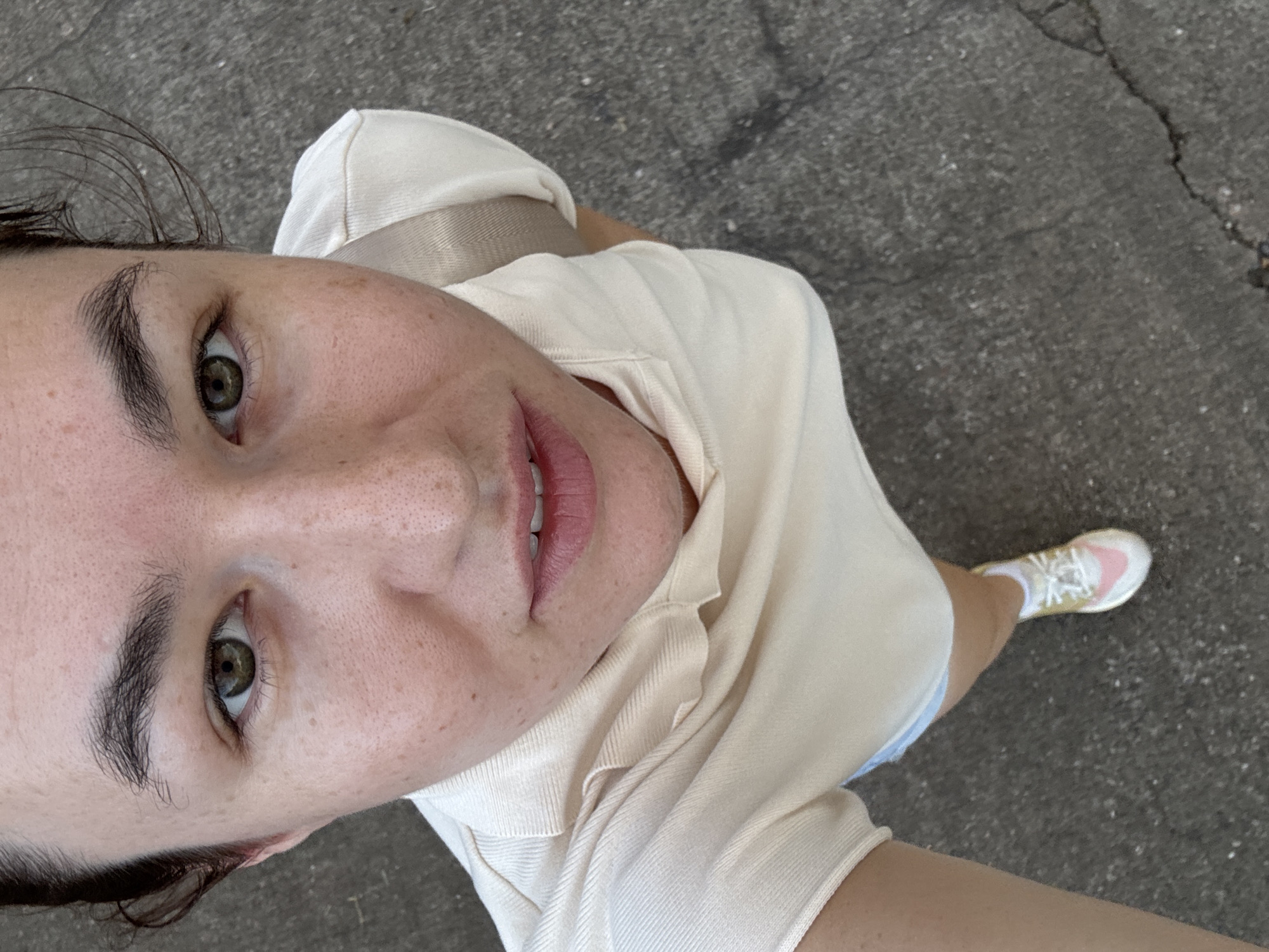 The image shows a close-up, high-angle shot of a woman's face and upper body. She has fair skin with freckles, dark eyebrows, and green eyes. She is wearing a light-colored, collared shirt and pink and white sneakers. Her hair appears to be dark and pulled back. The background is a cracked asphalt surface. No specific location within Surat Thani City Municipality, Thailand can be determined from the image.