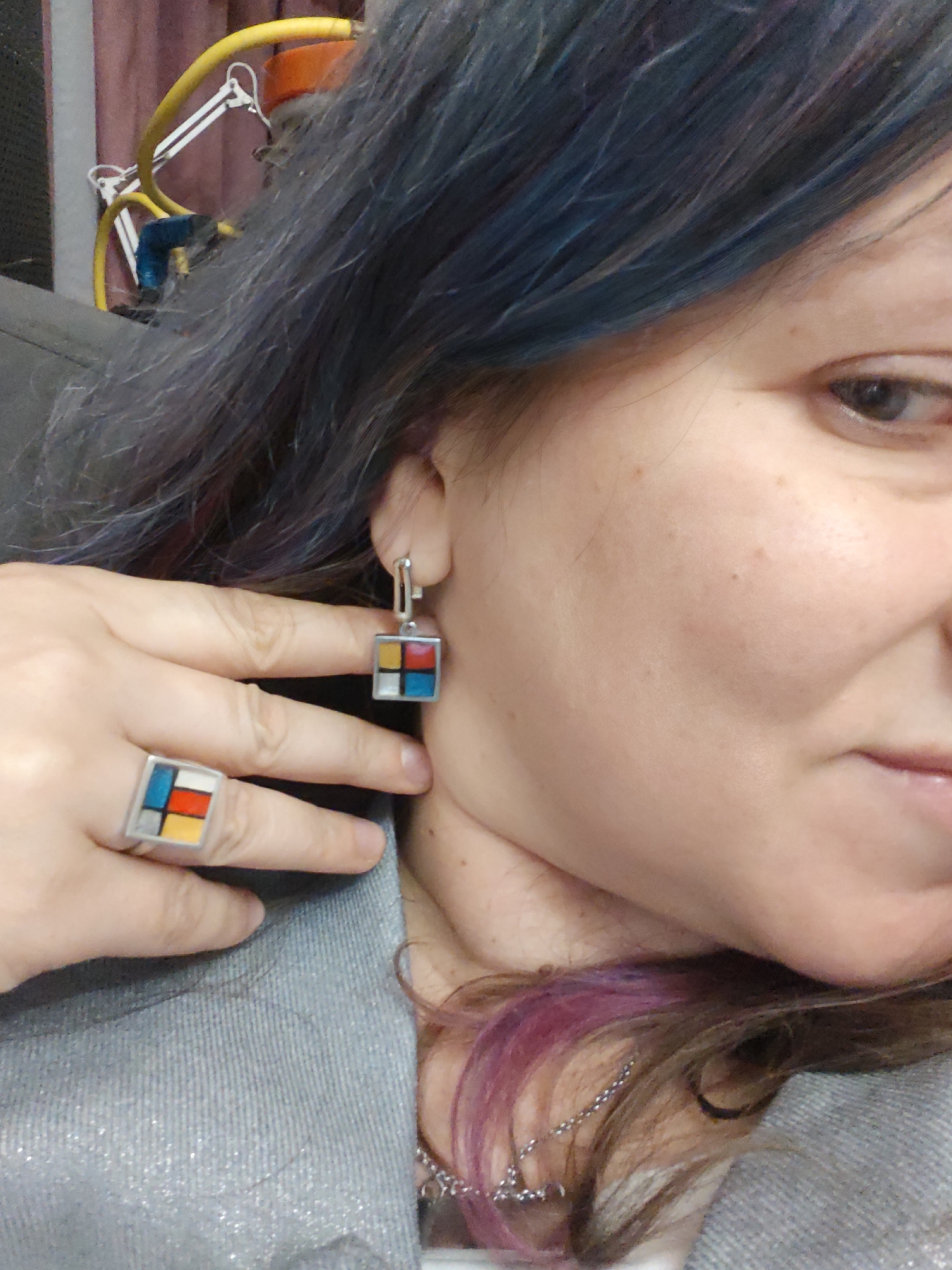 A close-up shot features a woman with blue-tinted hair wearing Mondrian-inspired jewelry, including a square ring and matching earrings. The jewelry is predominantly silver with small, colorful squares of red, yellow, blue, and white. The woman's hand is positioned near her ear, displaying the ring. She is wearing a gray, possibly metallic, shirt. The background includes a blurred yellow and white lamp and a glimpse of curtains. The location details (Свистуха, Russia) are not discernible from the image's content.