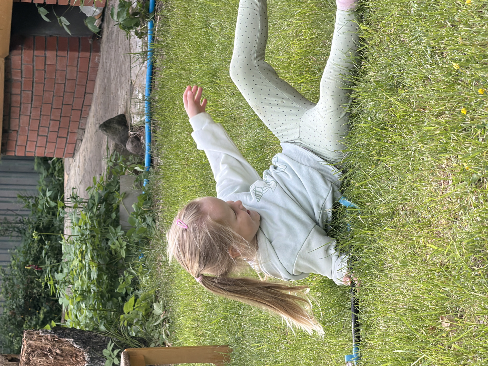 A child lies in green grass in an outdoor environment. The child wears light-colored leggings with dark polka dots and a light-colored sweatshirt. The child has blonde hair pulled back into a ponytail with a pink hair tie. The child's left arm is raised. Behind the child are brick buildings, foliage, and wooden structures. The location is likely Udelnaya, Russia.
