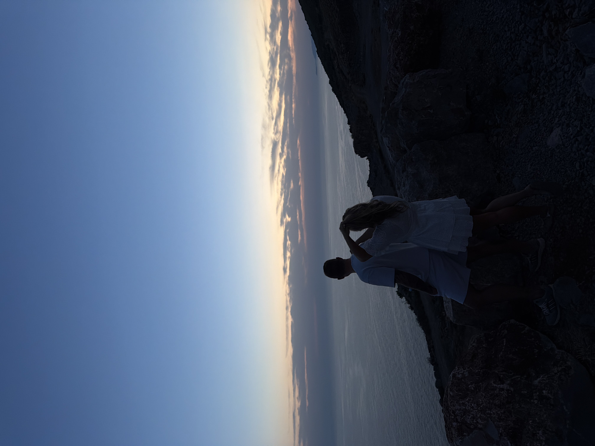 A silhouetted couple stands on a rocky outcrop overlooking the sea at dusk. The man, wearing a white t-shirt and shorts, stands with his back to the viewer, facing the ocean. The woman, in a white dress, stands next to him, with her hand raised to adjust her hair. The sky is a gradient of deep blue to light blue, with wispy clouds tinged with pink and orange from the setting sun. The sea is calm and reflects the colors of the sky. The rocky terrain is dark and textured, with large boulders in the foreground. The location is described as Kovačko Polje, Montenegro.