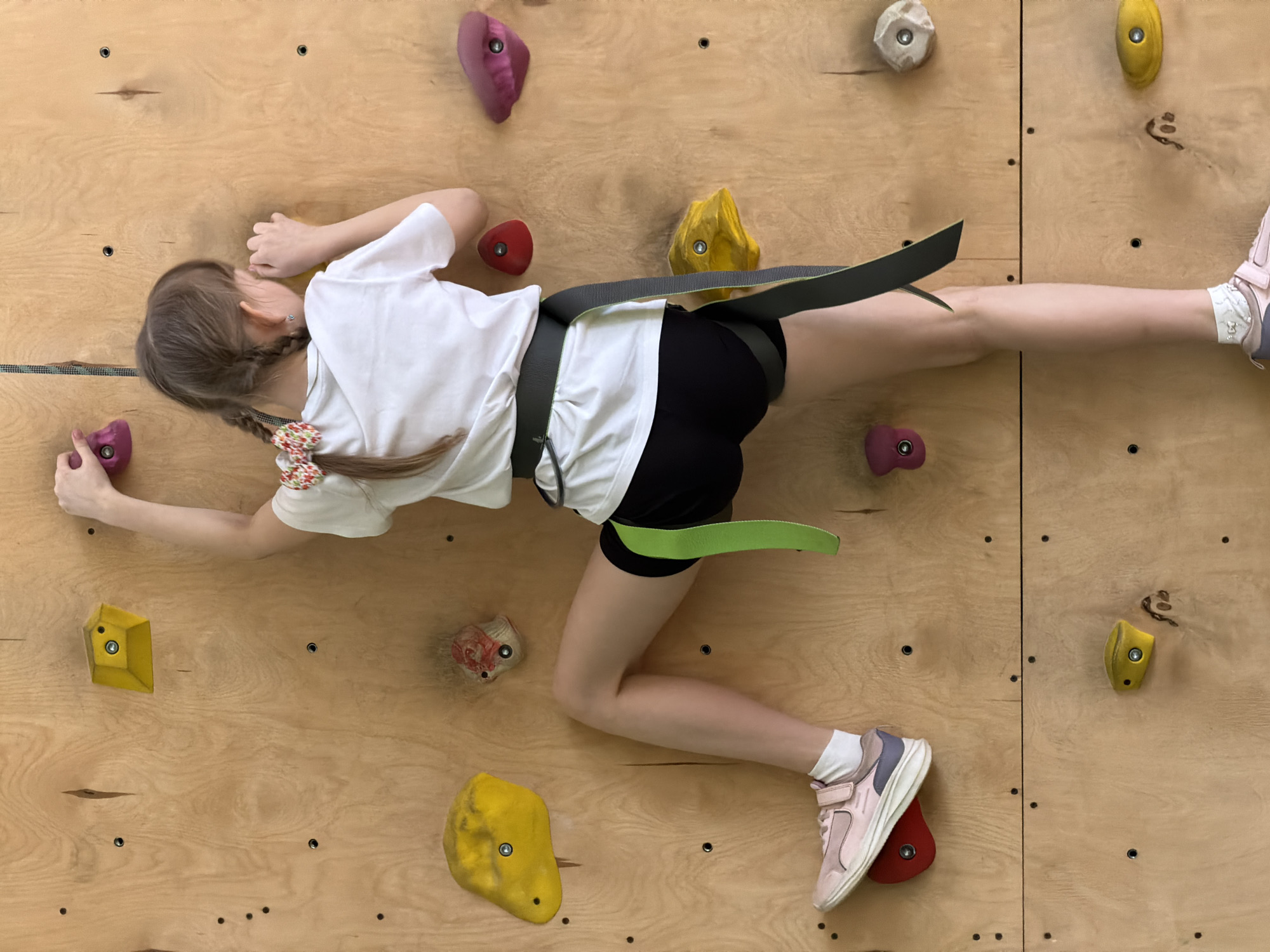 This image captures a young girl actively climbing or traversing an indoor climbing wall. She is positioned horizontally across light-colored wooden panels, her body stretched out as she uses various colorful climbing holds. Her left hand grasps a pink hold in the lower-left portion of the frame, while her left foot rests on a red hold. Her right hand is positioned near her head, and her right leg is bent.
The girl is dressed in a white t-shirt, black shorts, and light pink athletic shoes with white socks. A black and neon green safety belt or harness is secured around her waist and upper thighs. Her light brown hair is styled in two pigtail braids, each adorned with a colorful, floral-patterned scrunchie.
The climbing wall is constructed from large, light-toned plywood panels, featuring numerous screw holes for attaching the climbing holds. The holds themselves are varied in shape and color, including shades of pink/purple, yellow, red, and off-white/grey. The scene depicts a moment of physical activity, suggesting focus and effort during a climbing session. The setting is an indoor climbing facility, likely in Izhevsk, Russia. There is no visible text in the image.
