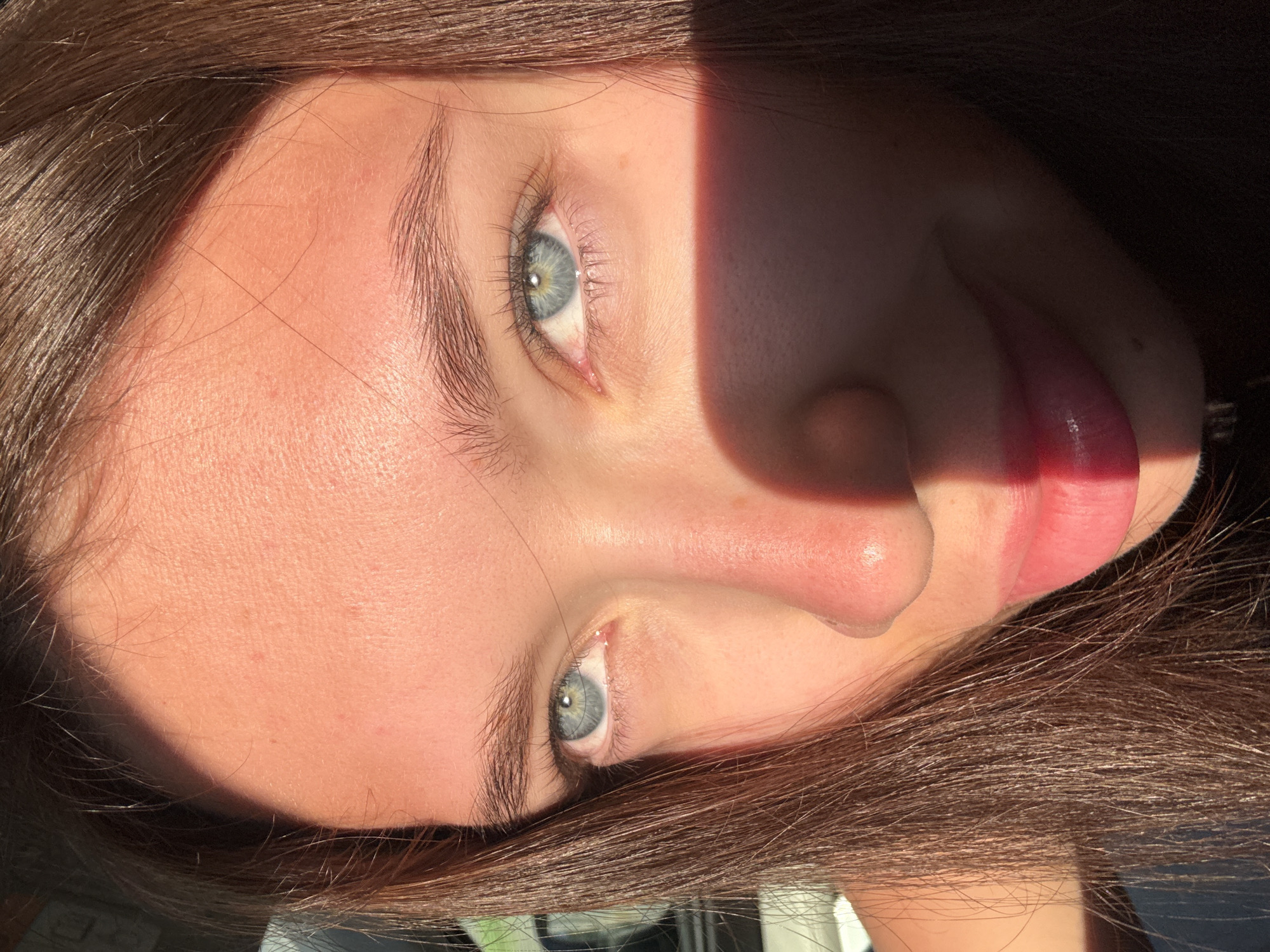 A close-up, rotated portrait shows a fair-skinned young woman with brown hair. The image is lit with harsh sunlight, creating sharp shadows on her face. She has light blue eyes, noticeable eyelashes, and defined eyebrows. Her nose is slightly prominent, and her lips are parted, revealing a hint of her tongue. The background is blurry and out of focus. The location and setting cannot be determined from the visual information provided.