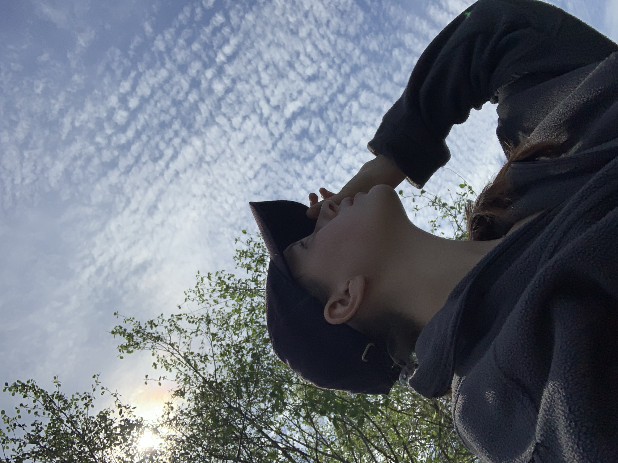 A low-angle, outdoor shot depicts a person wearing a dark baseball cap and a dark, textured jacket. The person is facing upward, their hand partially obscuring their face. The backdrop is a sky filled with altocumulus clouds. In the lower left corner, there are tree branches with green leaves. The sun is visible through the branches, creating a bright spot with a rainbow-like halo. There is no information in the image to indicate it was taken in Garevaya, Russia.