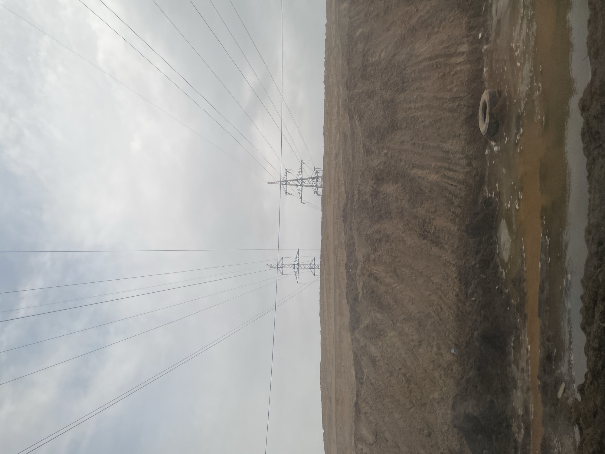The image depicts an outdoor scene near Kabanikha, Russia. In the foreground to the right of the frame, a dirt embankment borders a puddle of murky water. A discarded tire lies on the embankment. Overhead, power lines stretch across a cloudy sky, supported by two metal powerline towers in the middle-ground. The ground is devoid of vegetation.
