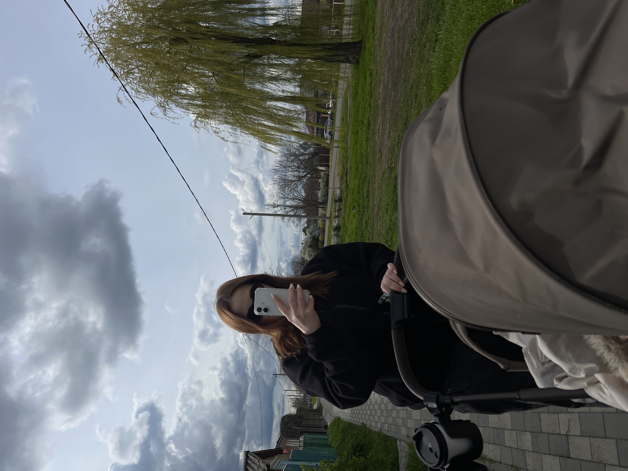 A person, possibly female with long auburn hair, is holding up a white smartphone with its camera facing them. They are wearing a black jacket and sunglasses. They are standing on a brick path next to a stroller. To the left is a small house with a turquoise exterior and an overgrown yard. Beyond the path is green grass, trees with minimal foliage, a telephone pole, and some houses. The sky is cloudy. The photo was taken in Novopokrovskaya, Russia.