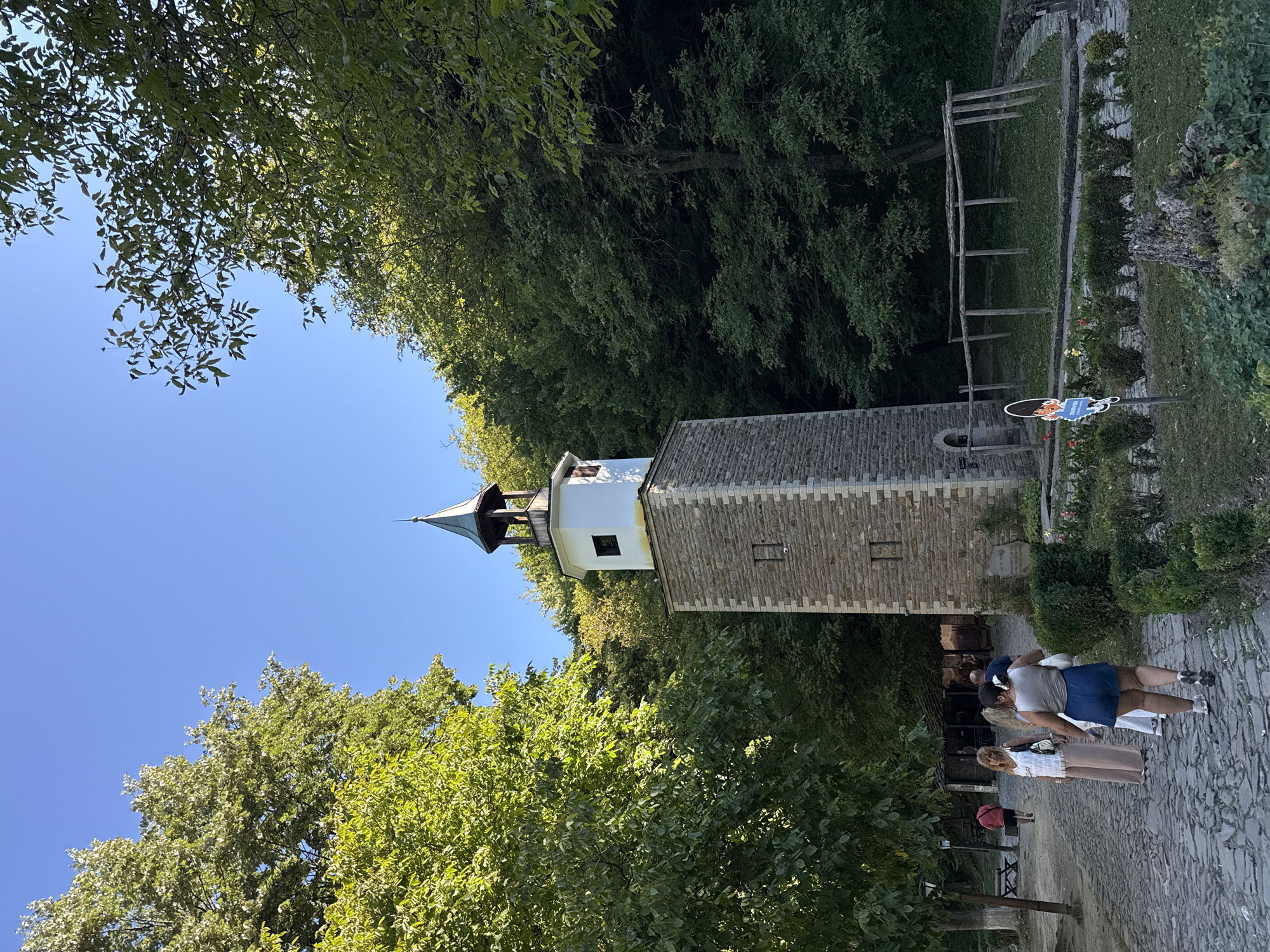 The image depicts a clock tower in the historical open-air museum of "Etar" in Gabrovo, Bulgaria. The tower, constructed of stone with a white upper section and a conical roof, stands tall amidst lush green trees. In the foreground, several people are walking on a cobblestone path. One woman in a white top and beige pants is walking away from the camera, while another woman in a light blue skirt and a grey tank top is also walking away. A third woman in a white dress is seen between them. Further in the background, a man in a red shirt walks away from the camera. The environment is natural, with abundant foliage surrounding the stone structures and pathways of the museum. The sky is clear and blue.