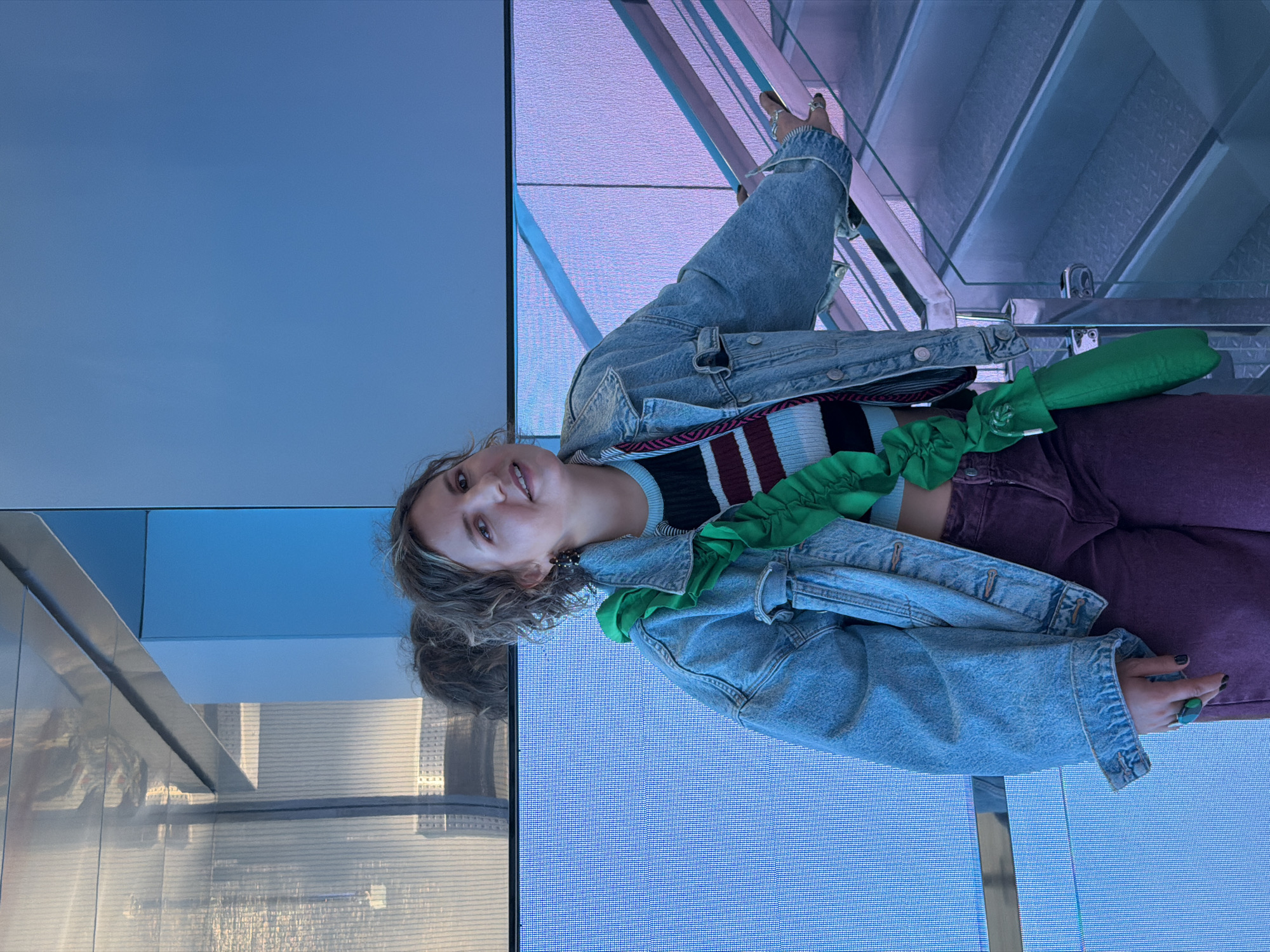 A fair-skinned female is positioned off-center in an upward-angled, brightly illuminated indoor shot. She is wearing a blue denim jacket over a striped top, with purple-hued pants. A green accessory is loosely draped across her midsection. The setting features a mix of glass and metal structures, including what appears to be a staircase with silver railings. The background includes a blue-toned wall and a view of what may be buildings. The image was captured within the Zaryadye Park media center in Moscow, Russia.