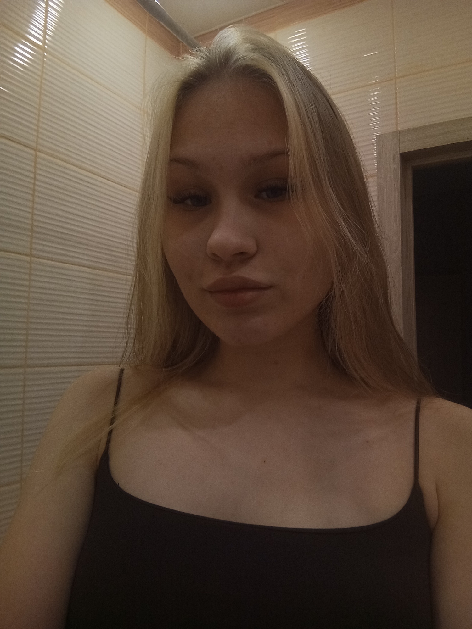 A young woman with light blonde hair with darker roots is posing for a selfie in what appears to be a bathroom. She is wearing a black tank top with thin straps. The background shows horizontally textured, light beige tile walls. Part of a doorframe and dark interior space is visible in the upper right corner. The image lacks identifying information about the city of Krasnodar, Russia, or any specific location within it. Therefore, a description of the location is impossible based solely on the image provided.