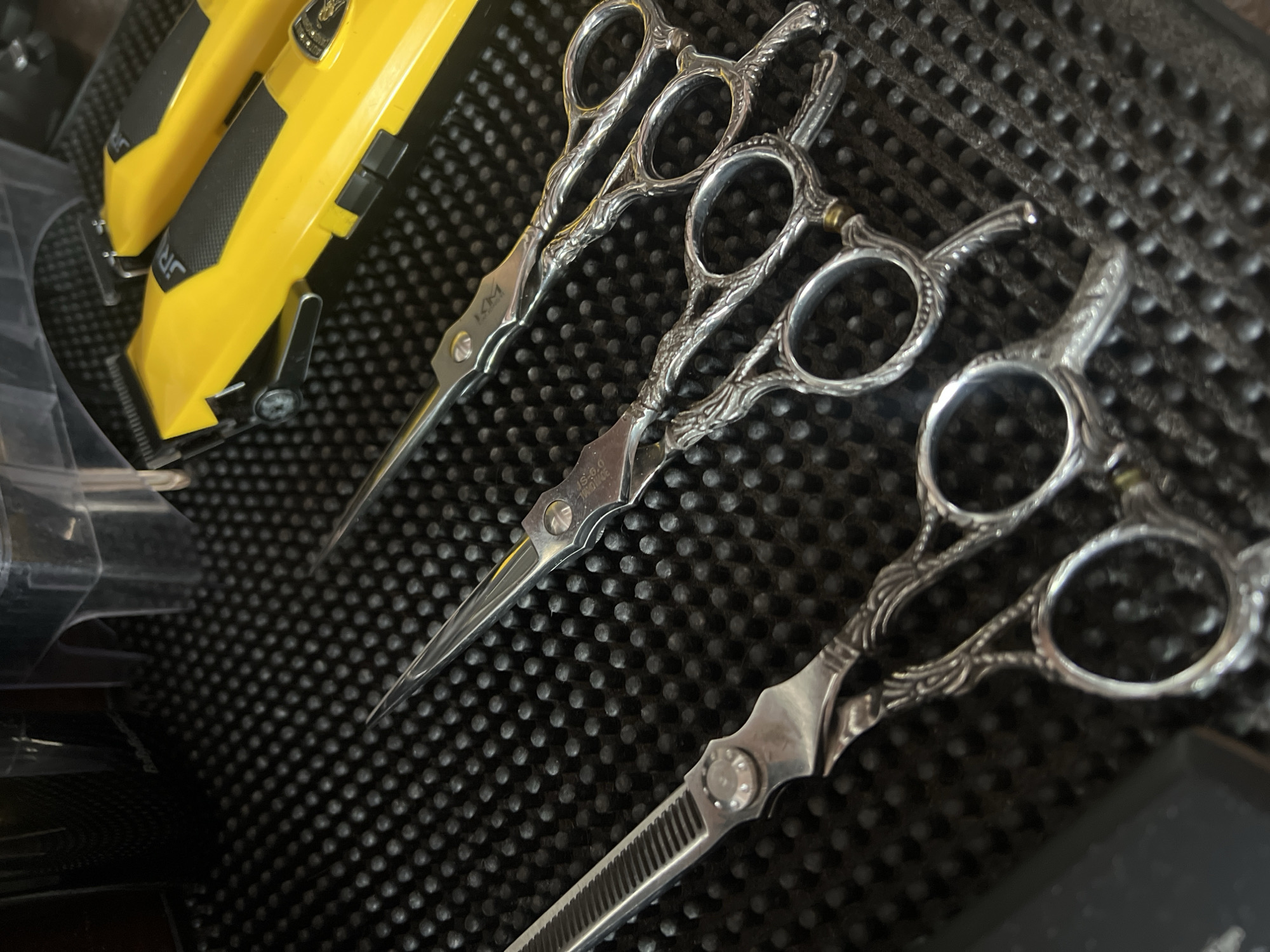 The image shows three silver-colored pairs of ornate hair-cutting shears or scissors arranged on a black, textured surface, possibly a mat. To the left, partially visible, are yellow and black hair clippers. The blades of the scissors are oriented towards the top of the frame. The handle designs are elaborate and feature circular finger holes. There is no information in the image to infer a location in Ternopil, Ukraine. No people, actions, or interactions are depicted.