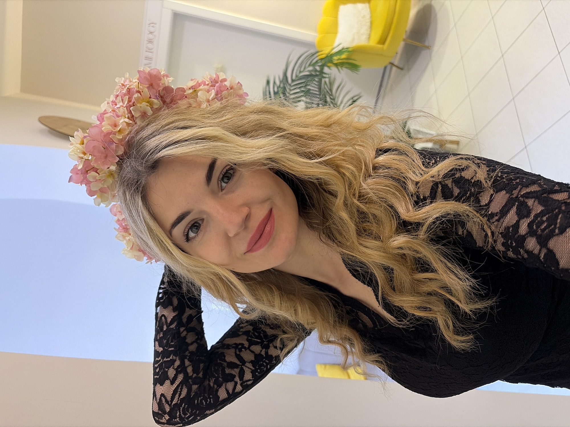 A woman with blonde, wavy hair wearing a flower crown of pink and white flowers is posing indoors. She is wearing a black lace top with long sleeves. The background includes a white wall with a white frame visible behind her, a yellow chair in the upper background, and a palm leaf. The lighting is bright and appears to be coming from overhead. The woman is looking directly at the camera with a slight smile. The location of the image is not discernable from the photo.