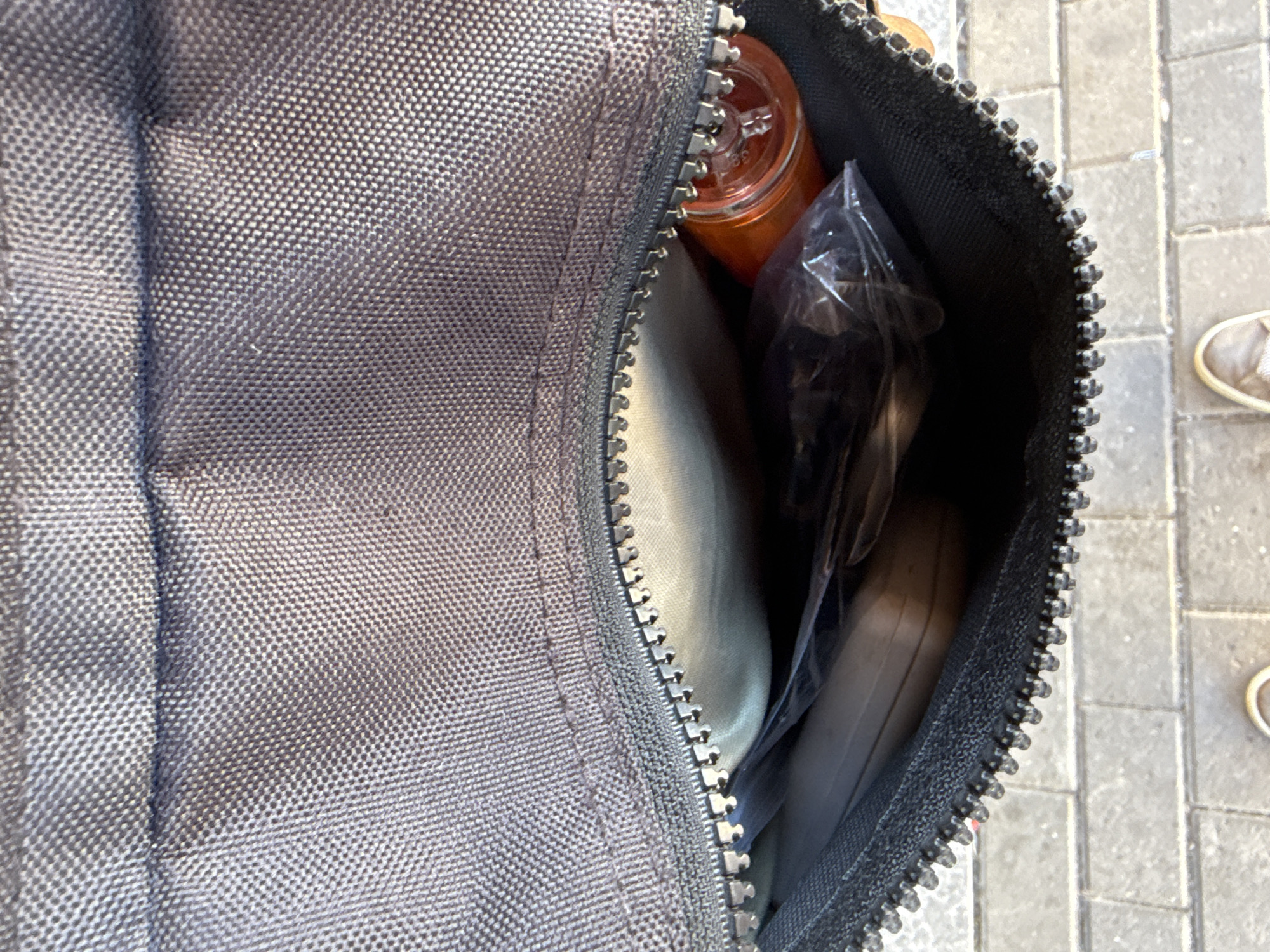 The image shows the inside of an open bag with a zipper. Inside the bag, there is a light-colored item, a clear plastic bag with dark items inside, and an orange bottle or jar with a metal lid. The bag appears to be made of a dark, textured fabric. Below the bag, a paved surface, possibly a sidewalk, is visible, with brick-like patterns. At the bottom of the image, two feet wearing light-colored shoes are visible, suggesting a person is standing or walking. The scene appears to be outdoors in an urban environment. There is no indication of the city of Bad Blankenburg, Germany.