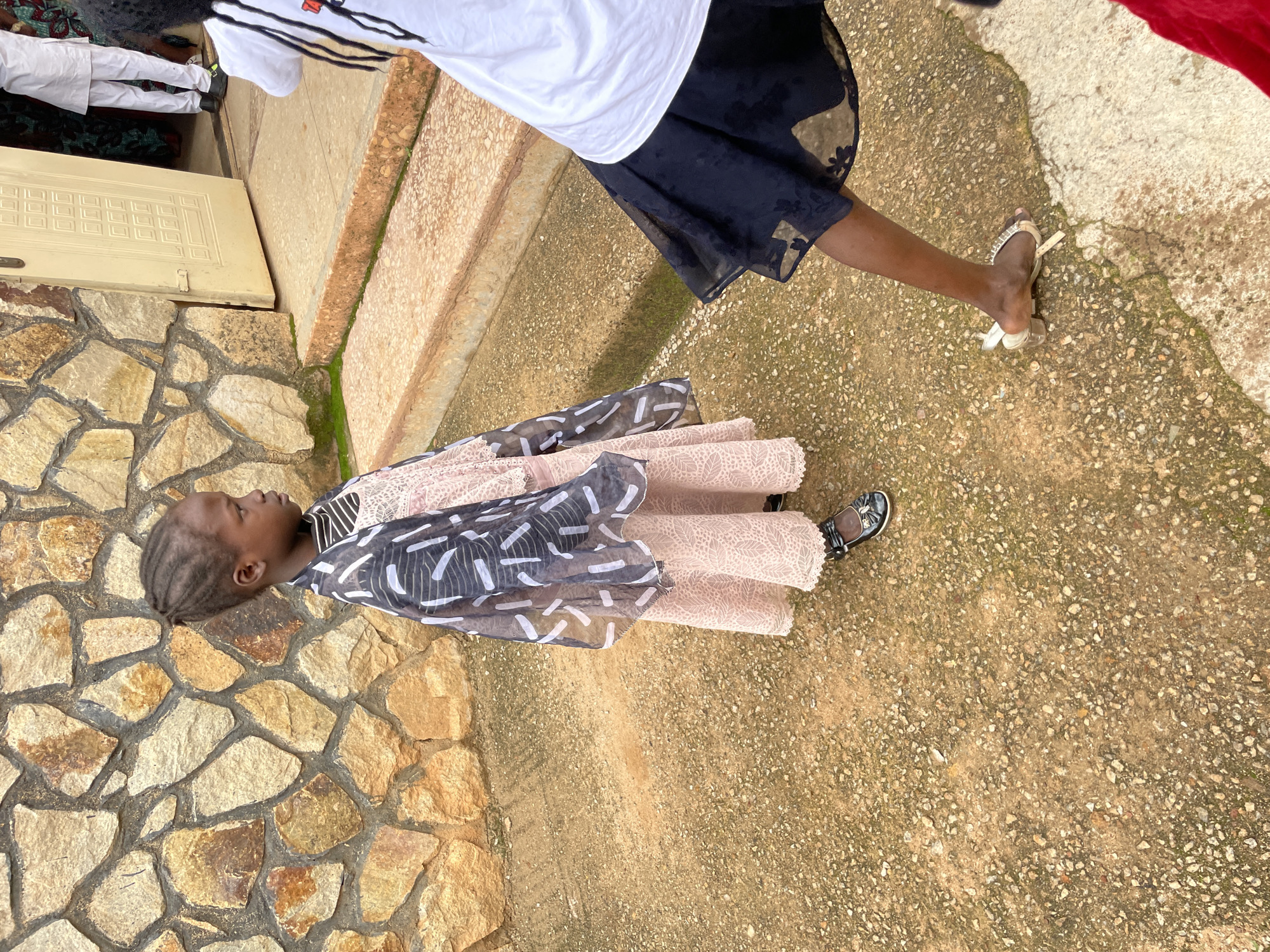 In Jos, Nigeria, a young girl stands near a stone-walled building with an open door. She wears a pink lace dress and a sheer, patterned overlay, accessorized with black Mary Jane shoes. To her right, another person, mostly out of frame, is visible from the waist down, wearing a black skirt and a white top. The ground is a mixture of concrete and gravel. The scene appears to be outdoors, possibly at a church or community center, judging by the architecture and the presence of people dressed for an event.