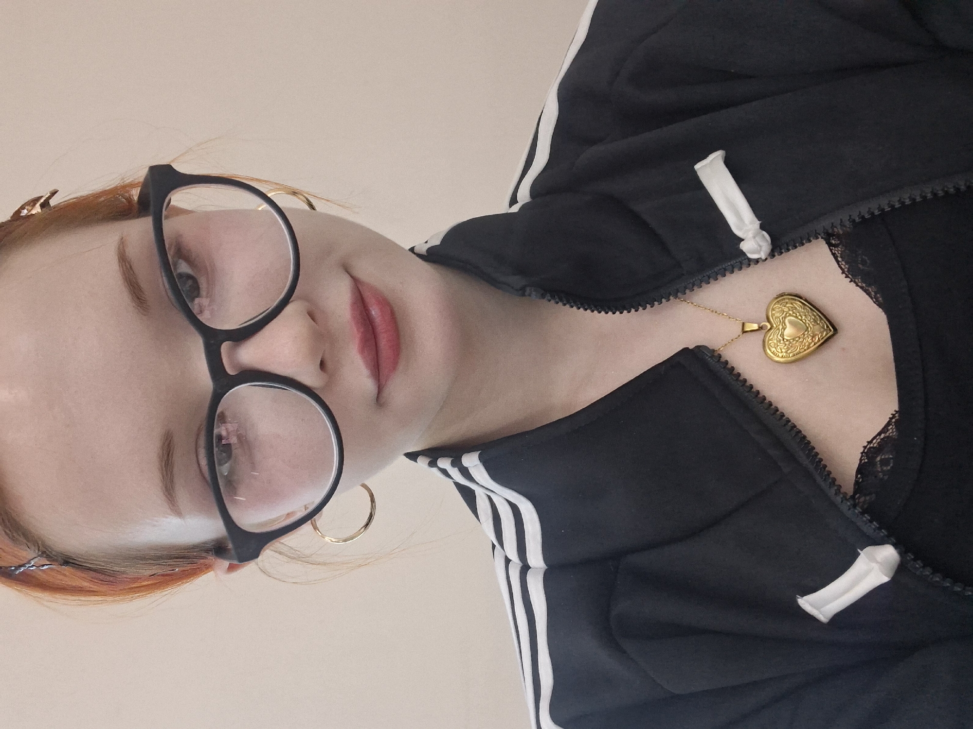 A close-up, tilted portrait of a fair-skinned person with red hair, wearing round black eyeglasses and gold hoop earrings. The person is wearing a black track jacket with three white stripes on the sleeves, partially zipped up to reveal a black lace top underneath. A gold heart-shaped locket necklace hangs around the person's neck. The background is a plain, light beige color. The photo does not provide information about the location in the city of Chaikovsky, Russia.