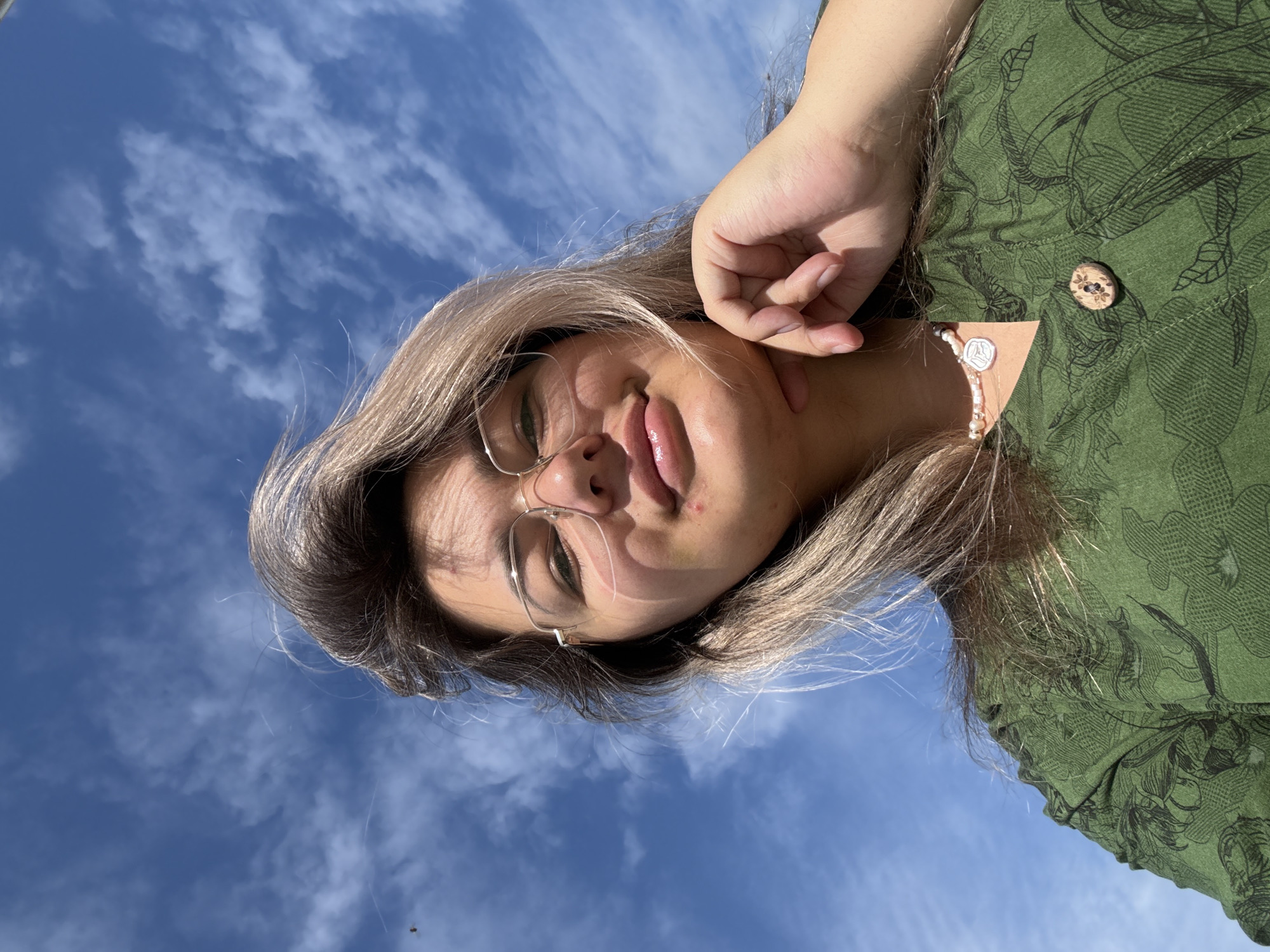 A young woman with light brown hair and glasses is captured from a low angle, looking up at a bright blue sky with scattered clouds. She is wearing a green patterned shirt with a wooden button. Her right hand is resting on her chin, and she is wearing a pearl necklace. The background is the sky, with a small dark object, possibly a bird, visible in the upper left corner. The overall lighting suggests it is daytime. The location is not specified in the image or its context.