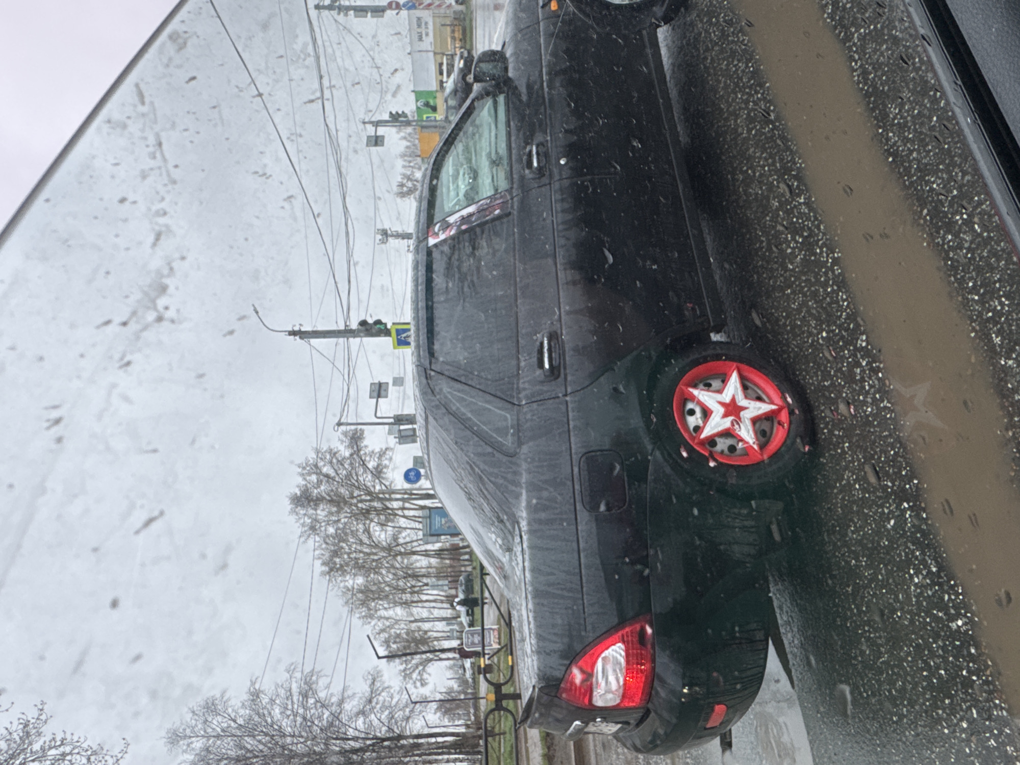 The image shows a black Lada Granta car parked on the side of a wet road in Chaikovsky, Russia. The car has a unique wheel cover featuring a red ring and a white star. The road is covered with water and dirt. The sky is overcast. In the background, there are leafless trees, utility poles with wires, and traffic lights. A pedestrian crossing sign is also visible.