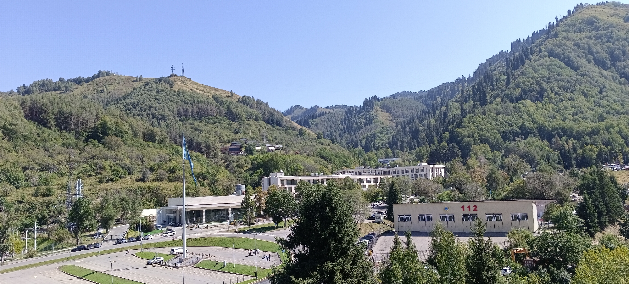 The image captures a panoramic view of a city district nestled at the foothills of a mountain range. In the foreground, a large paved area with parking spaces and a grassy median is visible. Several cars are parked in designated spots, and a few pedestrians are walking across the open space. To the left, a road stretches out, with traffic including cars and buses. A tall flagpole with a blue flag stands prominently near a modern, white, rectangular building. Further into the scene, another long, white building with multiple windows and floors is situated. To the right, a low-rise, beige building with several garage-like openings is marked with the large red number "112" and a circular emblem. The background is dominated by a lush, green mountain slope densely covered with trees, with power lines and transmission towers visible on the higher elevations. The sky is clear and blue. Based on the architecture, the flag, and the mountainous backdrop typical of the region, this scene is likely located in Almaty, Kazakhstan, possibly near the Institute of Engineering and Technology or a similar educational or administrative complex in the foothills of the Trans-Ili Alatau mountains.