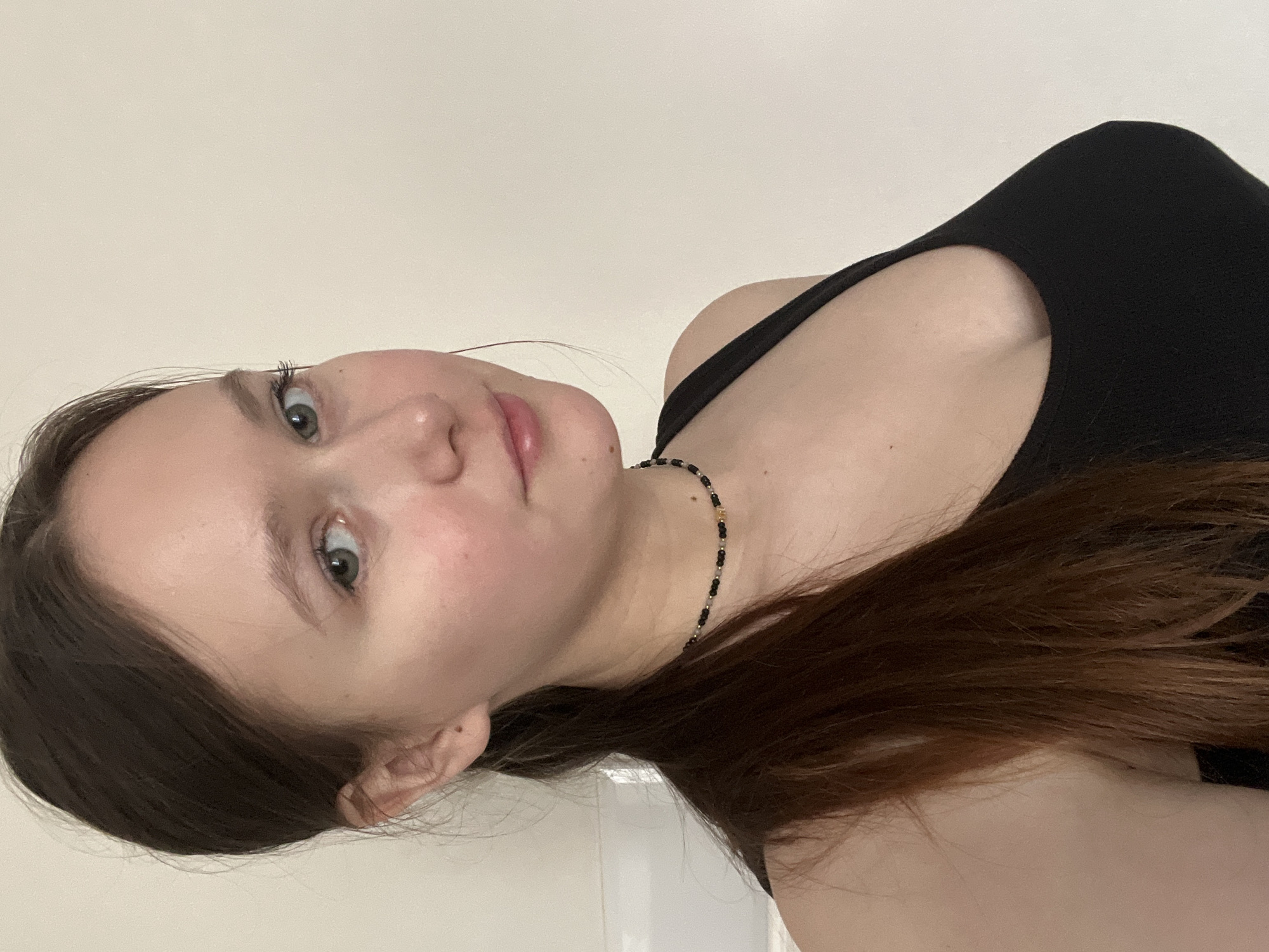 A portrait-style image, rotated 90 degrees counter-clockwise, features a young woman looking directly at the camera. Her head is positioned on the left side of the frame, with her body extending to the right. She has fair skin, light-colored eyes (appearing greenish-blue), and dark brown hair that falls over her shoulder and chest. Her lips are slightly parted, and she has a calm or neutral expression.
She is wearing a black tank top or camisole, with one thin strap visible over her right shoulder (as oriented in the image). A delicate necklace, composed of small black and gold beads, is visible around her neck. The background is a plain, light-colored wall, suggesting an indoor setting. The lighting is soft and even, highlighting her features without harsh shadows. There is no visible text in the image. The location context provided is Chaikovsky, Russia.