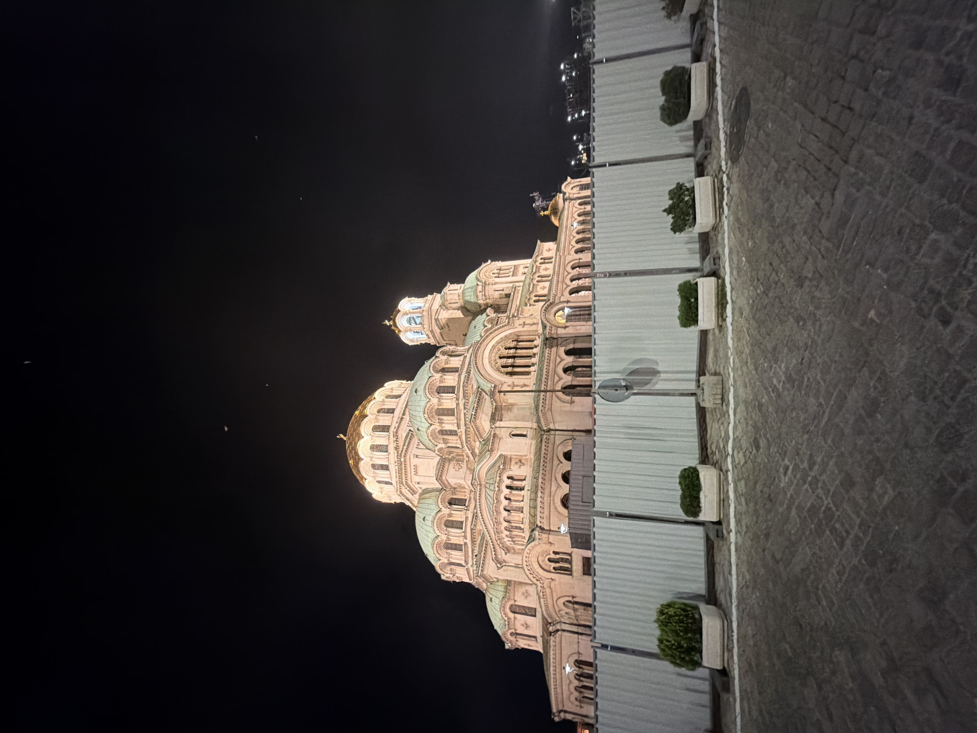 This is a nighttime, low-angle, wide-shot photograph of the Saint Alexander Nevsky Cathedral in Sofia, Bulgaria. The cathedral, a prominent landmark in the city center, is illuminated, standing out against a dark, starless sky.
The cathedral's cream-colored stone facade is brightly lit, showcasing its intricate architectural details. Multiple domes are visible; the largest central dome is gold, while several smaller surrounding domes are a muted greenish-teal. Crosses are visible atop the golden domes. Numerous arched windows line the various levels of the structure. On the far right, a bell tower with a light blue dome is visible.
In the immediate foreground, a temporary, light-colored metal fence, likely a construction or protective barrier, spans horizontally across the lower third of the image, partially obscuring the base of the cathedral. Behind the fence, several rectangular white planters containing small, dense green bushes are arranged along its length. A white line is painted on the dark, cobbled pavement in front of the fence. A thin pole with a circular sign or reflector is visible behind the fence on the right.
The pavement in the foreground is dark gray and appears to be made of cobblestones or pavers, exhibiting various textures and shades. The sky is deep black, with only a few faint, indistinct specks of light that might be distant stars or artifacts, and some distant bright lights visible on the far right horizon, possibly from a different urban area or event. No people are visible in the scene. The artificial lighting on the cathedral and foreground elements creates a strong contrast with the dark surroundings.
