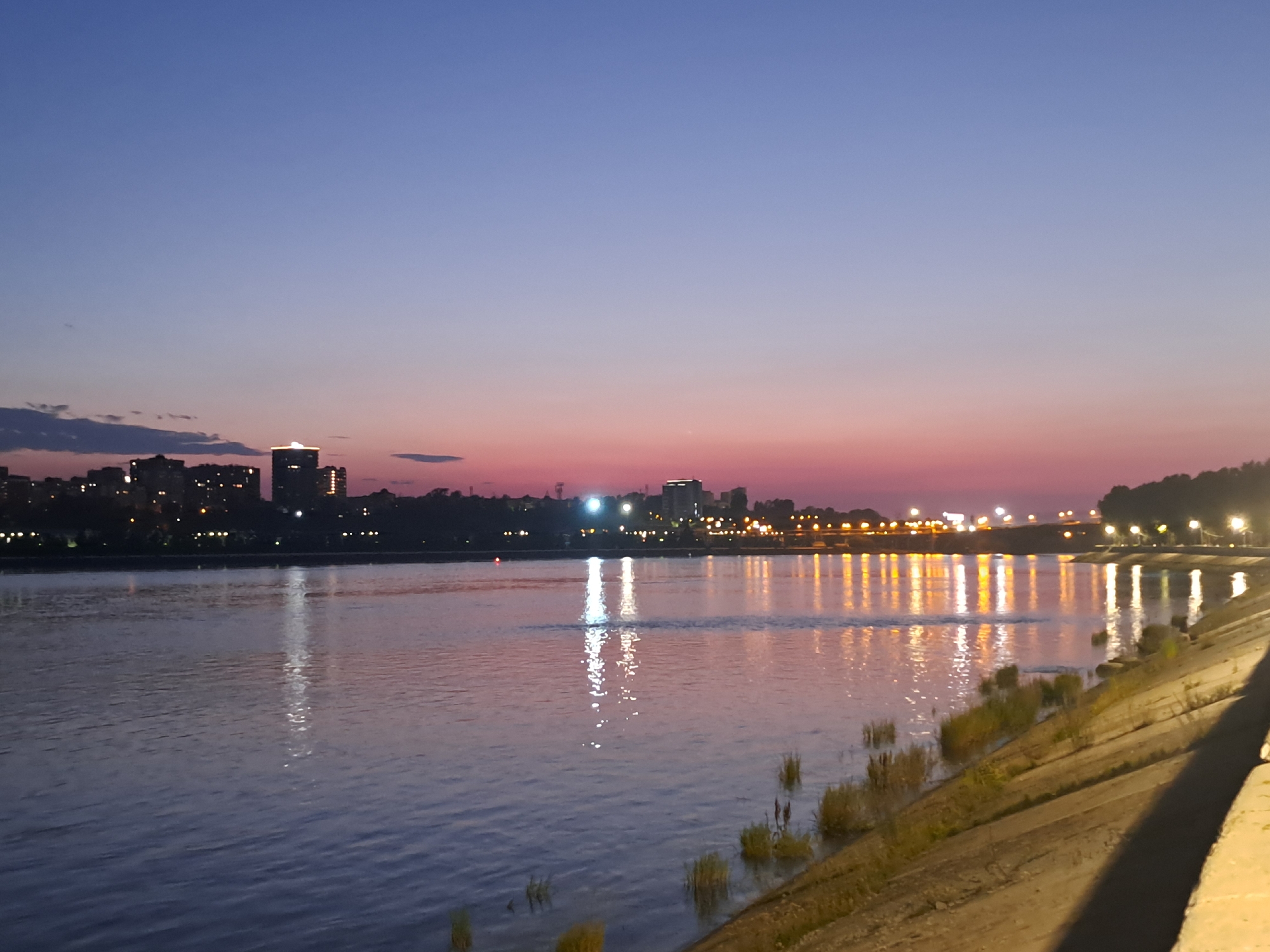 The image captures a serene twilight scene overlooking the Angara River in Irkutsk, Russia. The sky transitions from a deep blue at the top to a soft pink and orange hue near the horizon, indicating the sun has recently set. A city skyline, dotted with illuminated buildings and streetlights, stretches across the opposite bank of the river. The lights of the city are reflected in the calm water, creating a shimmering effect. The foreground features a concrete embankment sloping down to the river, with sparse vegetation growing along its edge. The overall atmosphere is peaceful and tranquil, showcasing the beauty of Irkutsk at dusk.