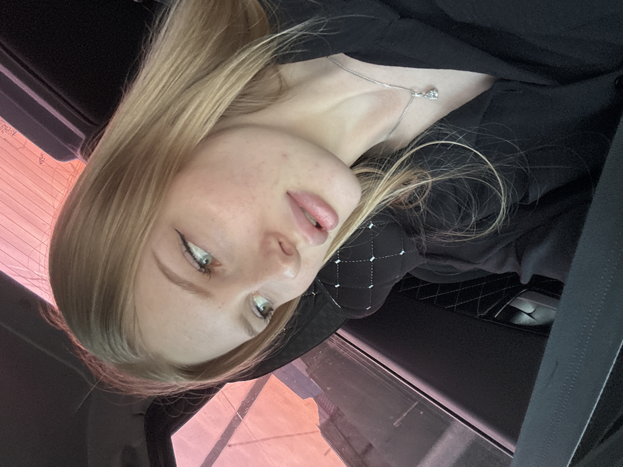 A young, fair-skinned woman with light blonde hair is seen in the foreground. She has freckles on her face and is wearing eyeliner. A delicate silver necklace adorns her neck. She is clad in a black garment. The setting appears to be inside a car, with a window visible in the background. The exterior view through the window reveals a building with pinkish hues. The seat behind her is black with a quilted pattern. The image offers no definitive clues to pinpoint the location as Кондратово, Russia, or any specific city for that matter.