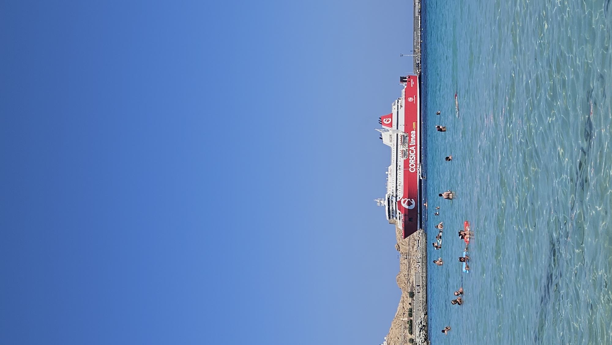 The image depicts a scenic view of a beach and the sea, with a large red and white ferry docked in the distance. The ferry, branded "CORSICA linea.com," is the prominent subject in the upper portion of the frame. The sea is clear and turquoise, with sunlight creating ripples and reflections on the surface. Several people are visible in the water, engaged in various activities such as swimming and floating on inflatable devices. The background features a rocky coastline with some buildings and vegetation. The sky is a clear, vibrant blue, indicating a sunny day. The scene evokes a vacation or leisure atmosphere, likely at a coastal town. While the specific location in L'Île-Rousse, France is not explicitly stated, the presence of the "CORSICA linea" ferry suggests a connection to Corsica, an island accessible from mainland France, and L'Île-Rousse is a port town on Corsica.