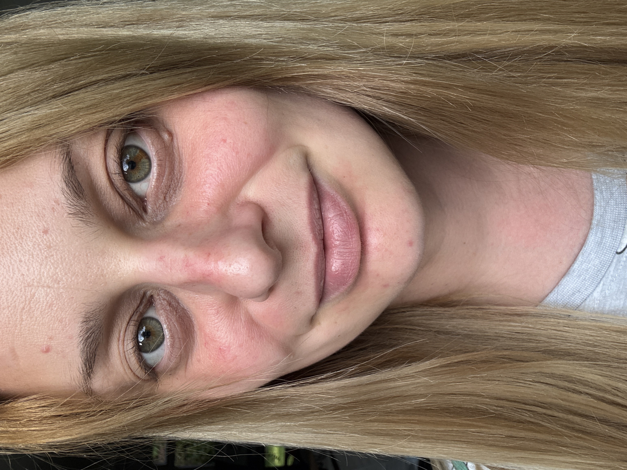 A fair-skinned woman with blonde hair is facing the camera with a slight smile. Her eyes are hazel in color. The background is blurry, hinting at a possible outdoor setting. The presence of a gray shirt at the periphery suggests she is wearing casual attire. There is no indication in the image that it is in Hazlemere, United Kingdom.