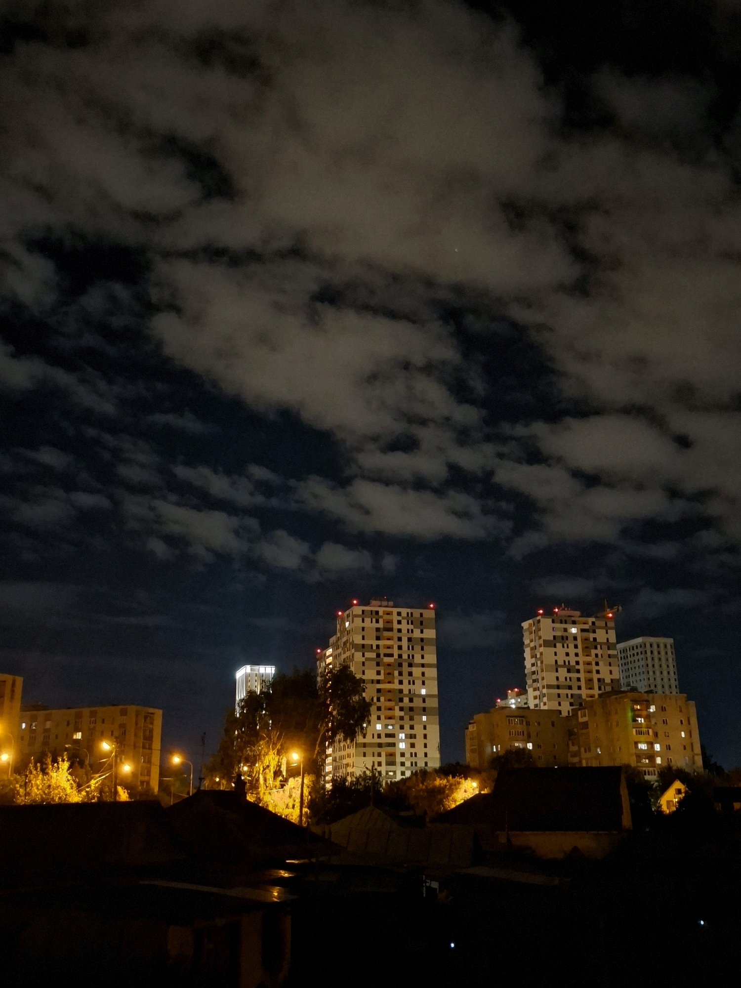 The image depicts a night scene in the city of Tyumen, Russia, featuring a cluster of apartment buildings against a cloudy sky. The buildings, some illuminated with interior lights and red warning lights on their upper levels, are of varying heights and architectural styles. The sky is filled with dramatic, dark clouds illuminated from below by the city lights, with a faint speck of light, possibly a planet or distant aircraft, visible in the upper center. In the foreground, the silhouettes of trees and lower structures are visible, with streetlights casting a warm glow on the surrounding foliage. The overall impression is of an urban landscape at night, with the natural elements of the sky adding a dynamic backdrop to the man-made structures.