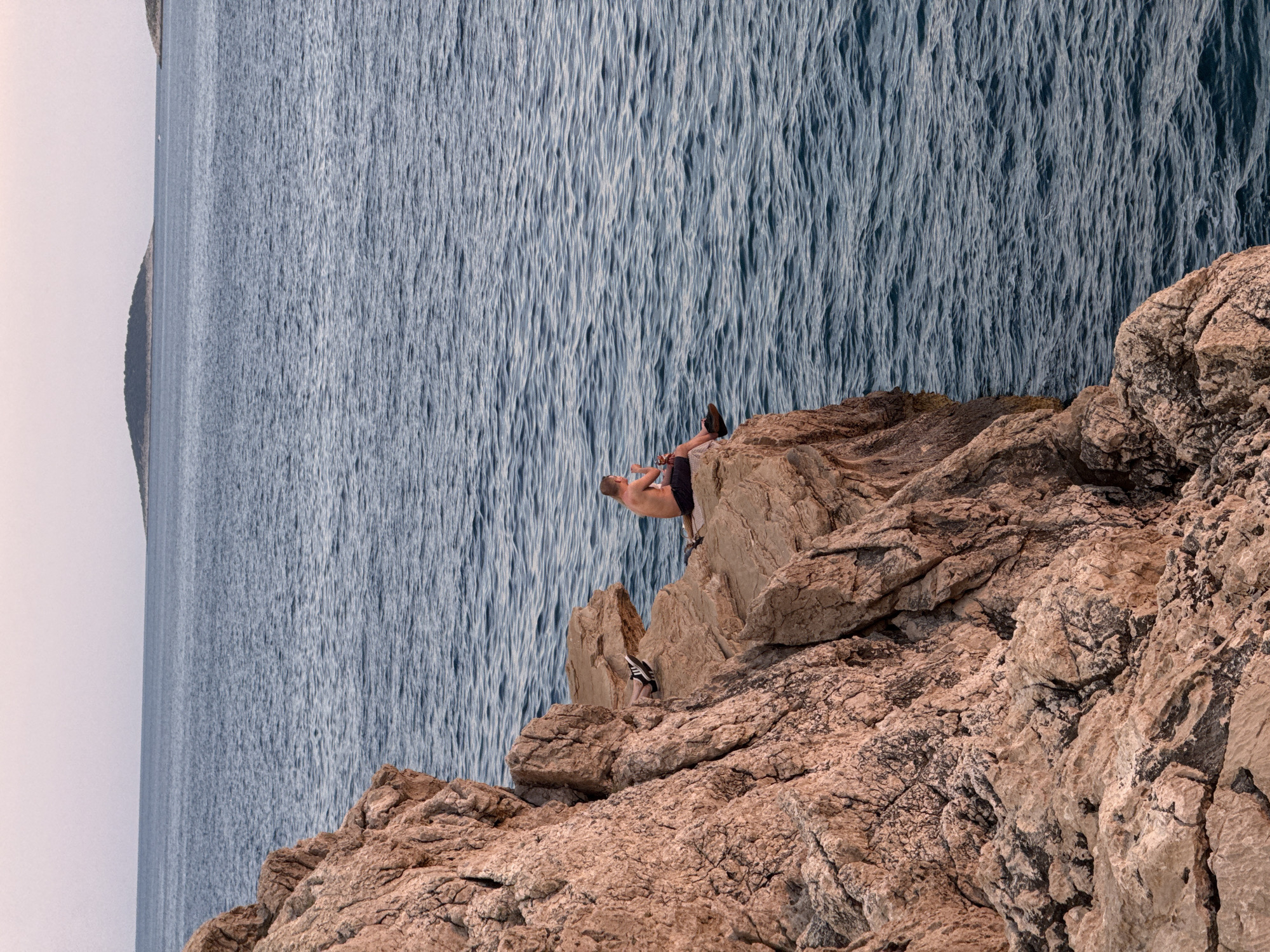 A young man, shirtless and wearing dark shorts, sits on the edge of a rocky cliff overlooking the Adriatic Sea in Primošten, Croatia. The cliff face, composed of rugged, reddish-brown rock, dominates the left side of the frame. The sea, a deep blue, stretches out to the horizon, where a few small, tree-covered islands are visible. The sky above is a pale, hazy pink, suggesting either dawn or dusk. The man appears to be holding a small object in his hands, and his gaze is directed out towards the water. His legs are casually crossed, with one foot resting on a lower ledge. The overall impression is one of peaceful solitude against a dramatic natural backdrop.