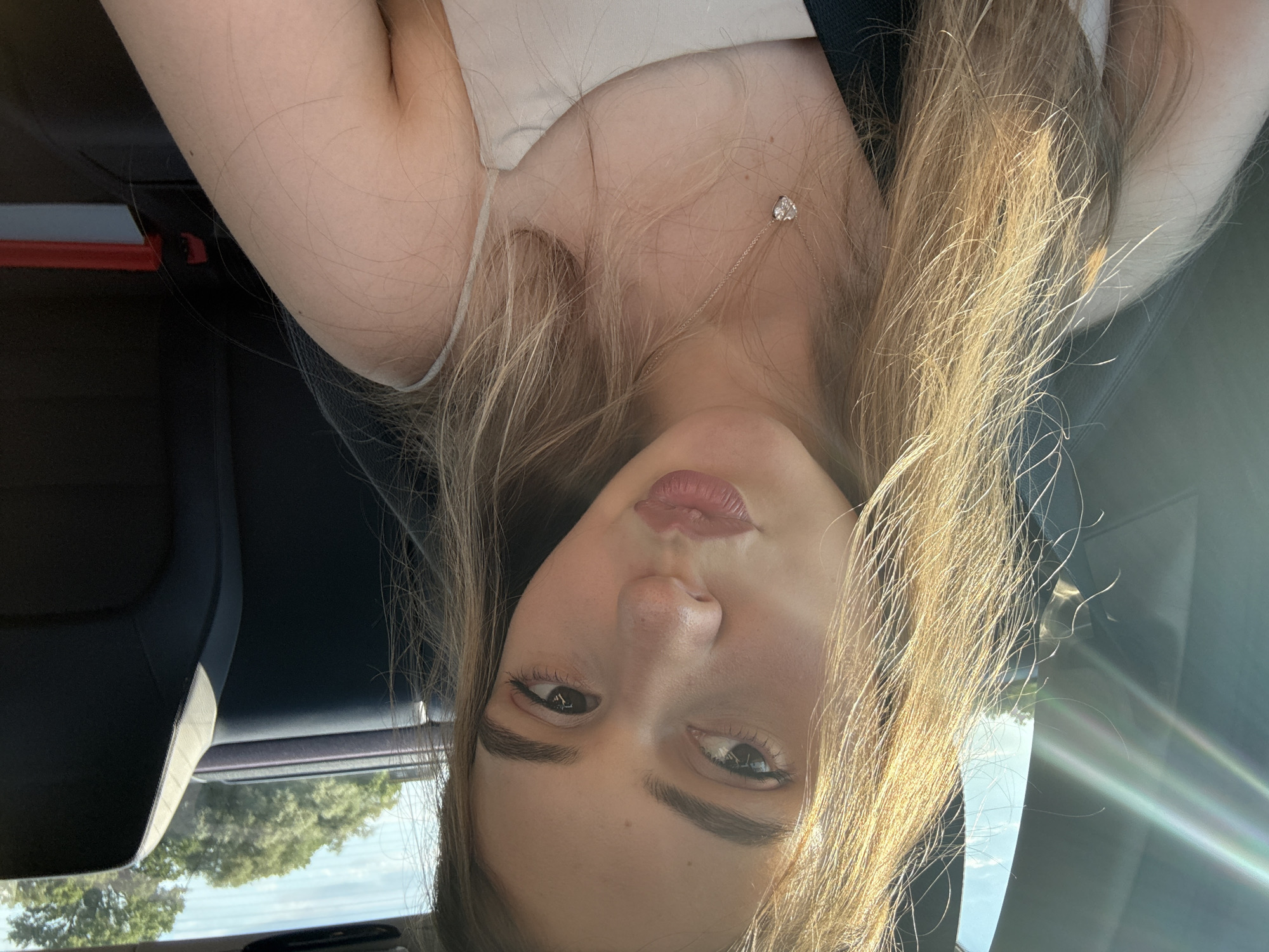 The media file shows a close-up, vertically oriented, selfie-style portrait of a young woman. She has long, blonde hair that is slightly windblown and catches the sunlight. She is wearing a thin-strapped top and a delicate necklace with a small heart-shaped pendant. Her face is turned towards the camera, with her eyes looking directly at the viewer. She has on subtle makeup, including eyeliner and lip gloss. The background is dark and appears to be the interior of a car, possibly a seat and a headrest. Sunlight is streaming in from the right side, creating lens flares and illuminating her hair. Outside the car window, through a slightly blurred view, a bright sky and green trees can be seen, suggesting it is daytime with fair weather. The overall mood is casual and relaxed, possibly taken while traveling. There is no visible text in the media file.