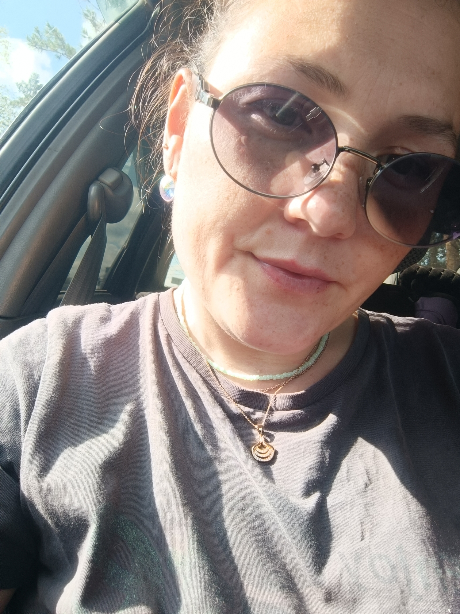 The image shows a close-up, selfie-style shot of a woman wearing a gray t-shirt and two necklaces, one with green beads and another with a pendant. She is also wearing round sunglasses with a subtle purple tint and a single stud earring in her left ear. The woman is in the passenger seat of a car, as indicated by the seatbelt buckle and car door visible in the background. Sunlight is streaming through the window, casting shadows on her face and shirt. The woman's skin has visible freckles. The image does not contain any information about the city of Foki, Russia.