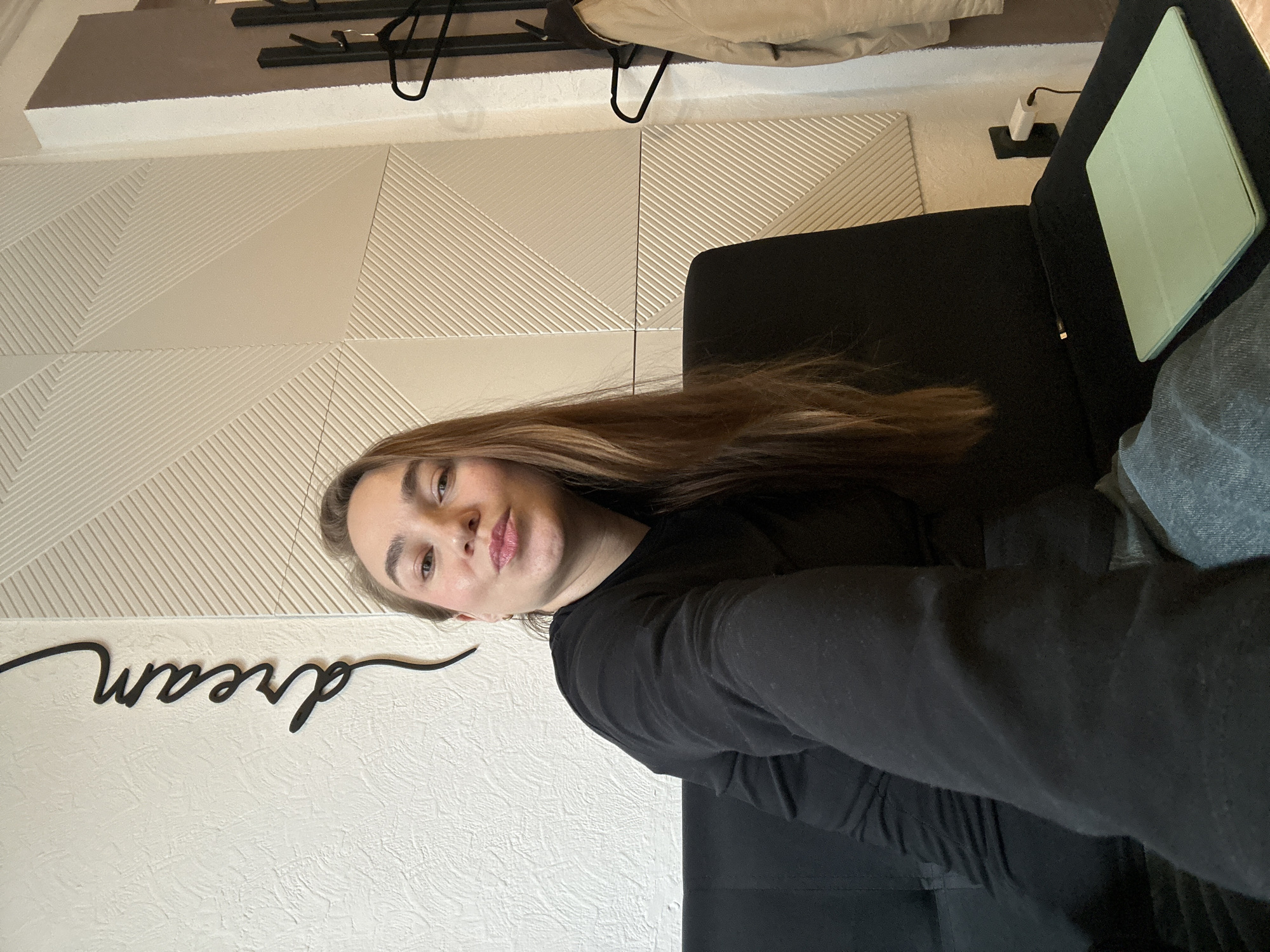 A woman is lying on a dark-colored couch, wearing a black shirt. The couch is next to a white wall with a black "dream" sign. Above the wall is a light-colored patterned wall and a coat rack with a beige coat. To the right of the woman is a closed tablet in a light green case. The location is Chaikovsky, Russia.