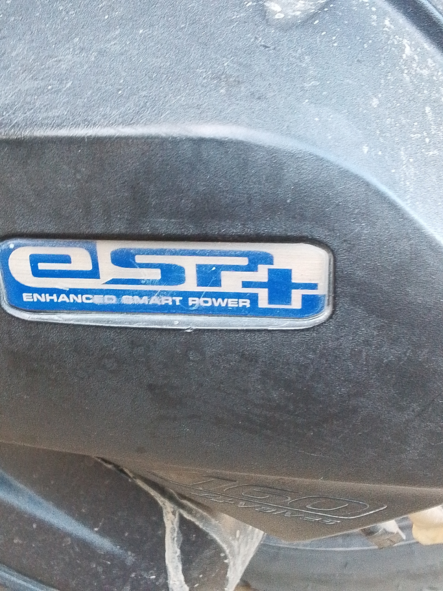 The image shows a close-up of the "eSP+" (Enhanced Smart Power) logo on a vehicle, potentially a scooter or motorcycle. The logo is silver with blue accents and the words "ENHANCED SMART POWER" are printed beneath the "eSP+" abbreviation. The logo is affixed to a dark grey, textured surface. The lower portion of the image reveals a partial view of the vehicle's body, with some white residue or dirt visible. The inscription "160" is faintly discernible near the bottom edge. The setting is unknown and the location in Lhokseumawe, Indonesia cannot be verified from the image. No people, actions or interactions are visible.
