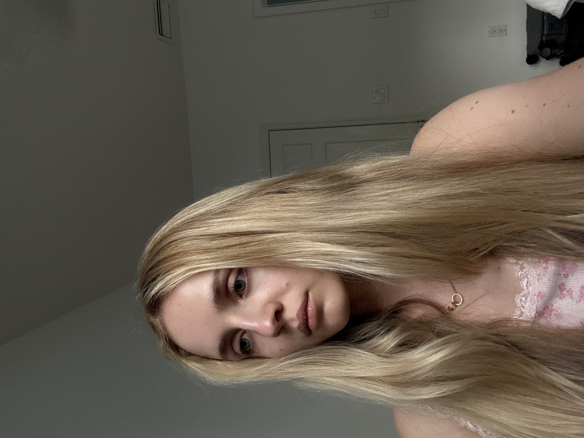 A close-up, low-angle, portrait-oriented photograph shows a young person with long, blonde hair draped over their shoulder and chest. They are looking directly at the camera with a neutral expression. The subject is wearing a light-colored, possibly pink, top with lace detailing and a floral pattern. A delicate gold necklace with a small pendant is visible around their neck.
The background is dimly lit and appears to be an interior setting. A grey wall occupies most of the left side of the frame, with a doorway and what looks like a light switch or electrical outlet visible on the right side. The lighting suggests it might be daytime, but the indoor setting and dim light make it difficult to determine the exact time. The overall mood of the image is calm and introspective. There is no visible text in the image.