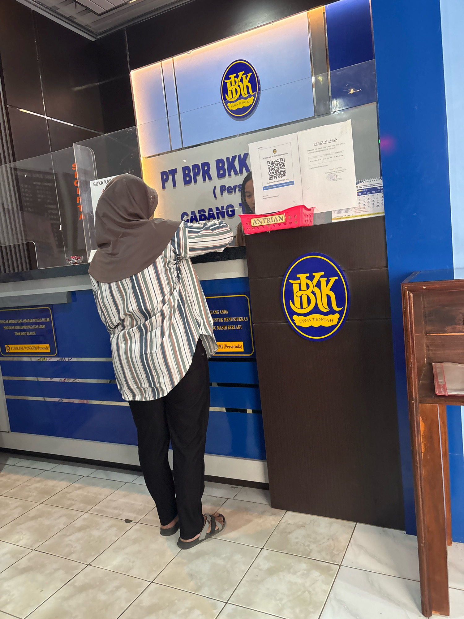 The image depicts the interior of a BPR Bank branch, likely in Tirtomoyo, Indonesia, given the text on the signage. A woman wearing a hijab and a striped long-sleeved shirt is standing at a counter, interacting with a teller who is partially visible. The counter has a glass partition for safety. Behind the counter, there is signage with the bank's logo and name, "PT BPR BKK". There are also various documents, a pink basket labeled "ANTRIAN" (queue/waiting line), and a calendar visible on the counter. The bank's interior features blue and dark brown paneling. The floor is tiled with a light-colored pattern. To the right of the counter, a wooden furniture piece is visible. The lighting suggests an indoor environment, possibly during business hours.