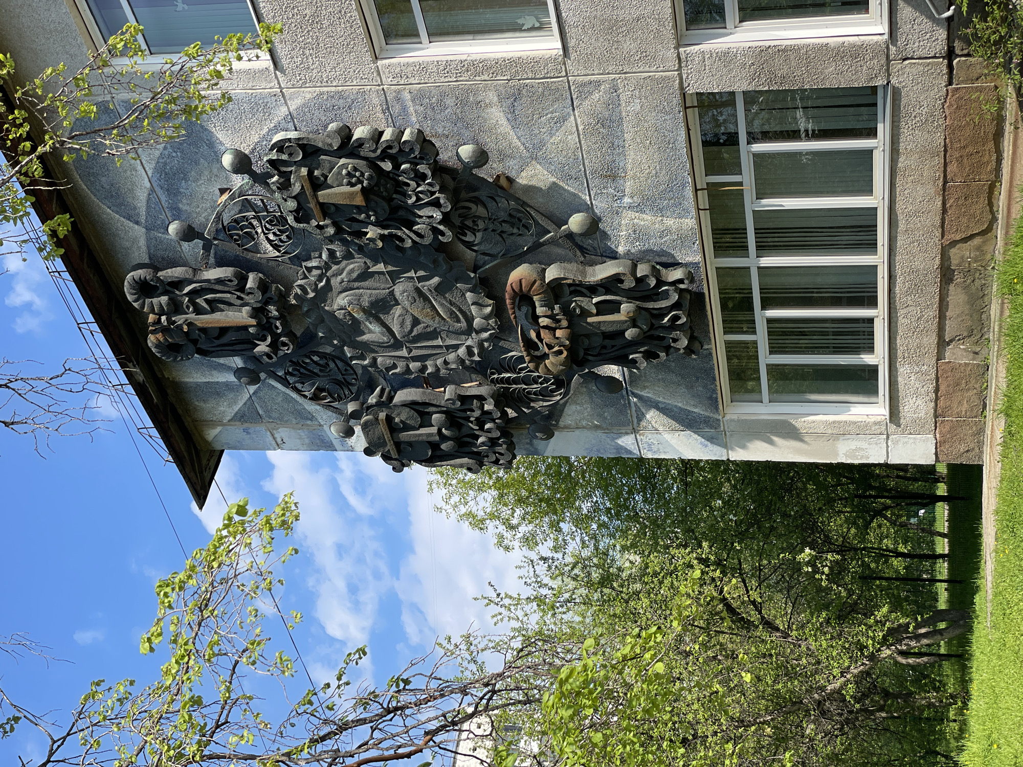 The image depicts a building in Zheleznogorsk, Russia, featuring a decorative metal relief mounted on its facade. The building has rectangular windows with white frames. The relief, composed of dark metal elements, is complex and abstract, possibly depicting industrial motifs or symbolic figures. Trees with green foliage are visible in the foreground, alongside a patch of grass. The sky is blue with scattered clouds.