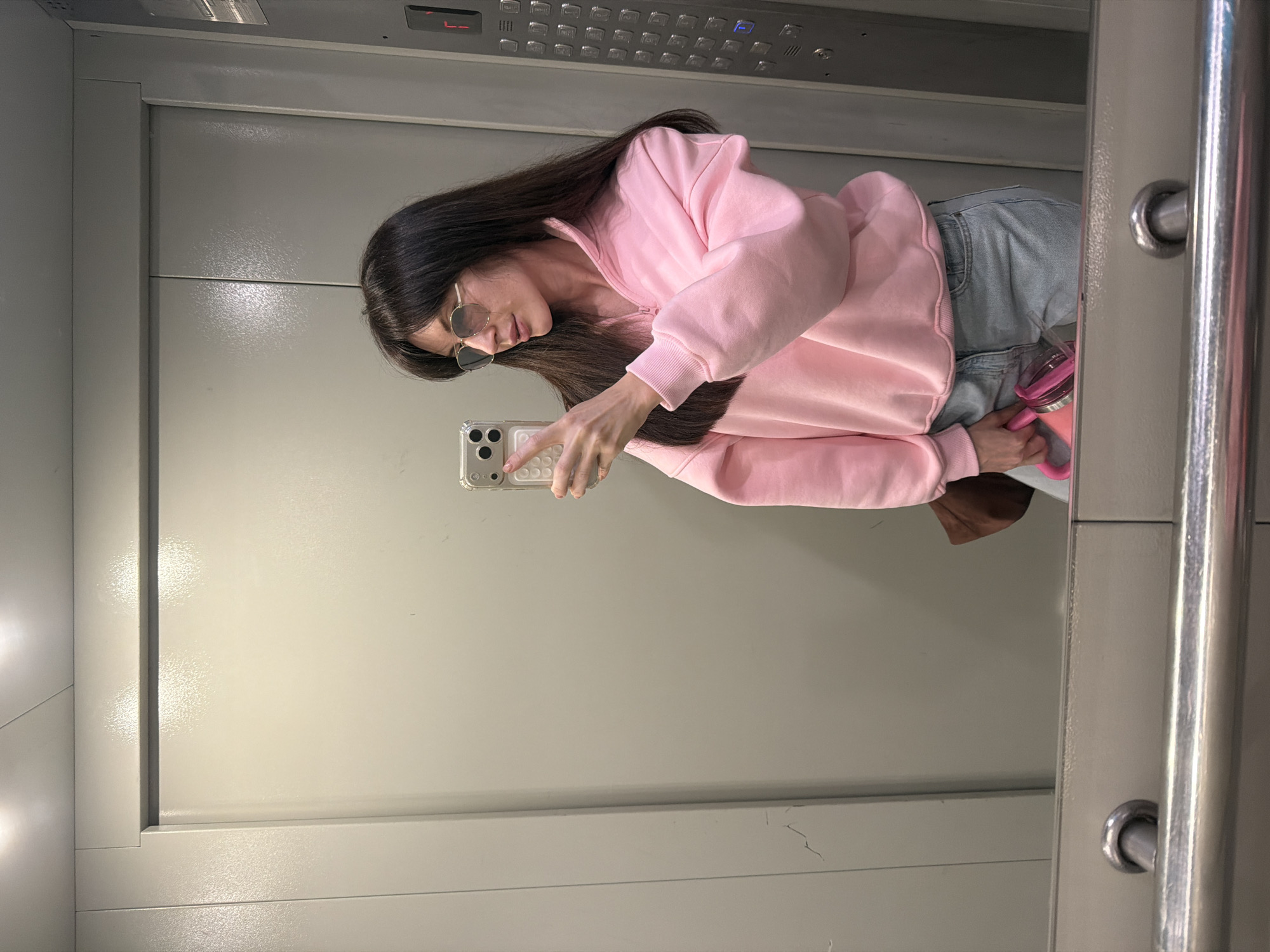 A person with dark hair and sunglasses is taking a selfie in an elevator with a smartphone. The person is wearing a pink sweatshirt and jeans and holding a pink water bottle. The elevator's control panel is visible, along with the door. The location is Perm, Russia.
