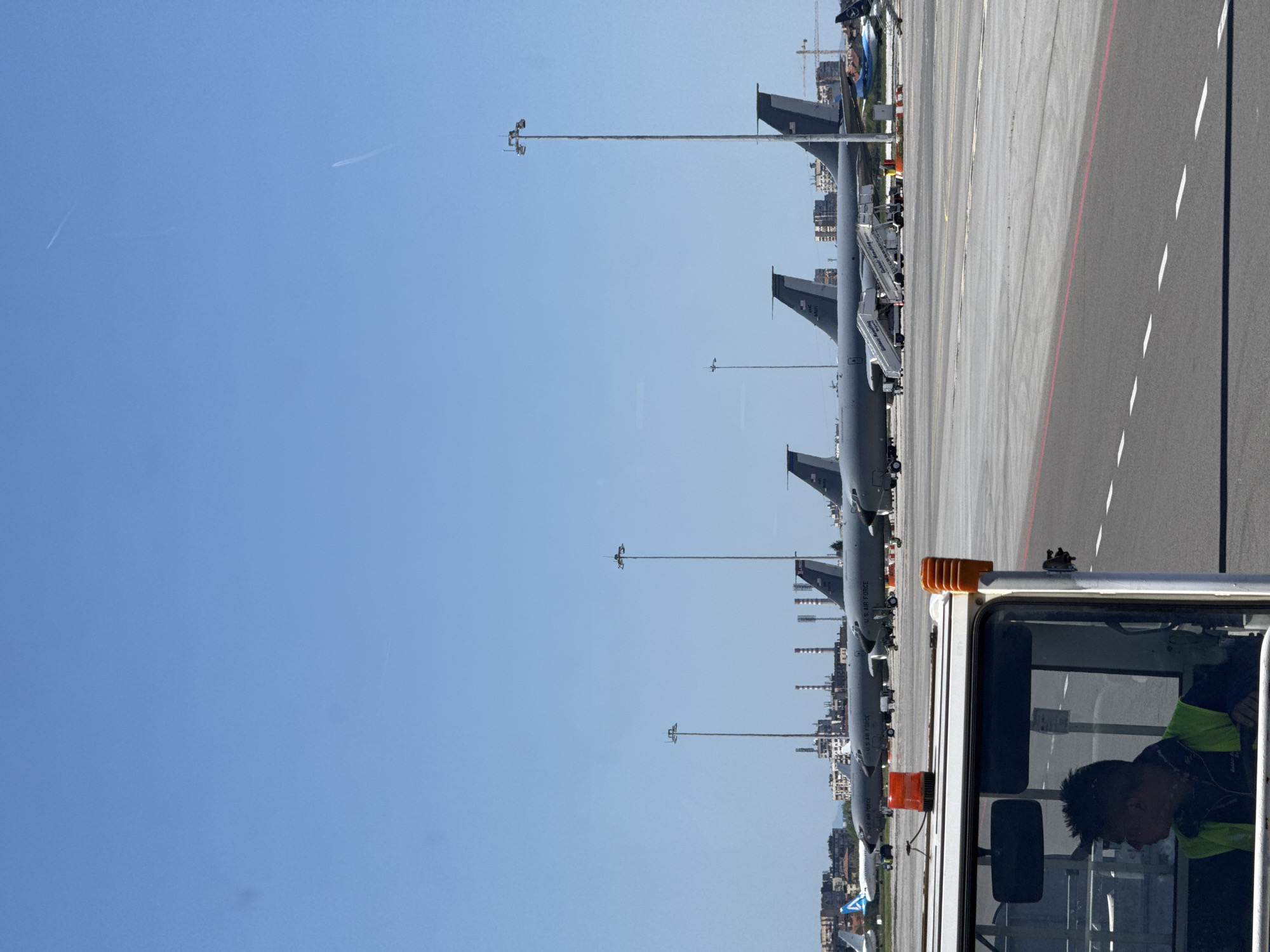 The image captures a row of military aircraft, specifically KC-135 Stratotankers, at Sofia Airport in Bulgaria. The aircraft are parked on the tarmac with service vehicles nearby. In the foreground, there is a utility vehicle with a person inside, possibly a ground crew member. The background shows elements of Sofia's urban landscape. The sky is clear and blue.