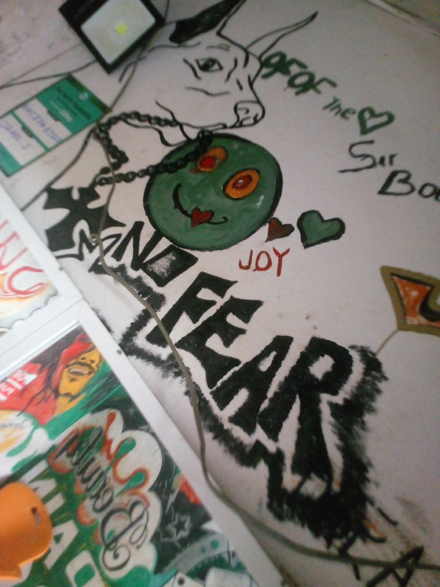 This media file captures an array of artistic drawings, stickers, and framed prints displayed on a light-colored wall, likely indoors. The main subject is a large, hand-drawn illustration featuring a cartoonish bull's head in the upper left, connected by a drawn chain to a circular green face with red and orange eyes, resembling a stylized smiley.
Prominently displayed below the green face is the large, stylized text "NO FEAR" in black with white outlines, emanating a sense of boldness. Adjacent to this, in red, is the word "JOY," accompanied by a small red heart and a green heart. Near the bull's head, green stylized text reads "gsof the <3 Sir Boo".
Other visible elements include a small black rectangular light fixture with a white LED panel at the top. Below this are two green rectangular cards or stickers, possibly identification cards, though their text is largely blurred. On the left side, partially visible, are framed pictures; one depicts a person's face (reminiscent of a Rasta figure like Bob Marley) in vibrant colors, and another showcases abstract, graffiti-style lettering. At the bottom left, an orange fish-shaped object is partially visible, possibly a small toy or decoration. The overall scene suggests a creatively decorated personal space. No specific activity is taking place; it's a static display.