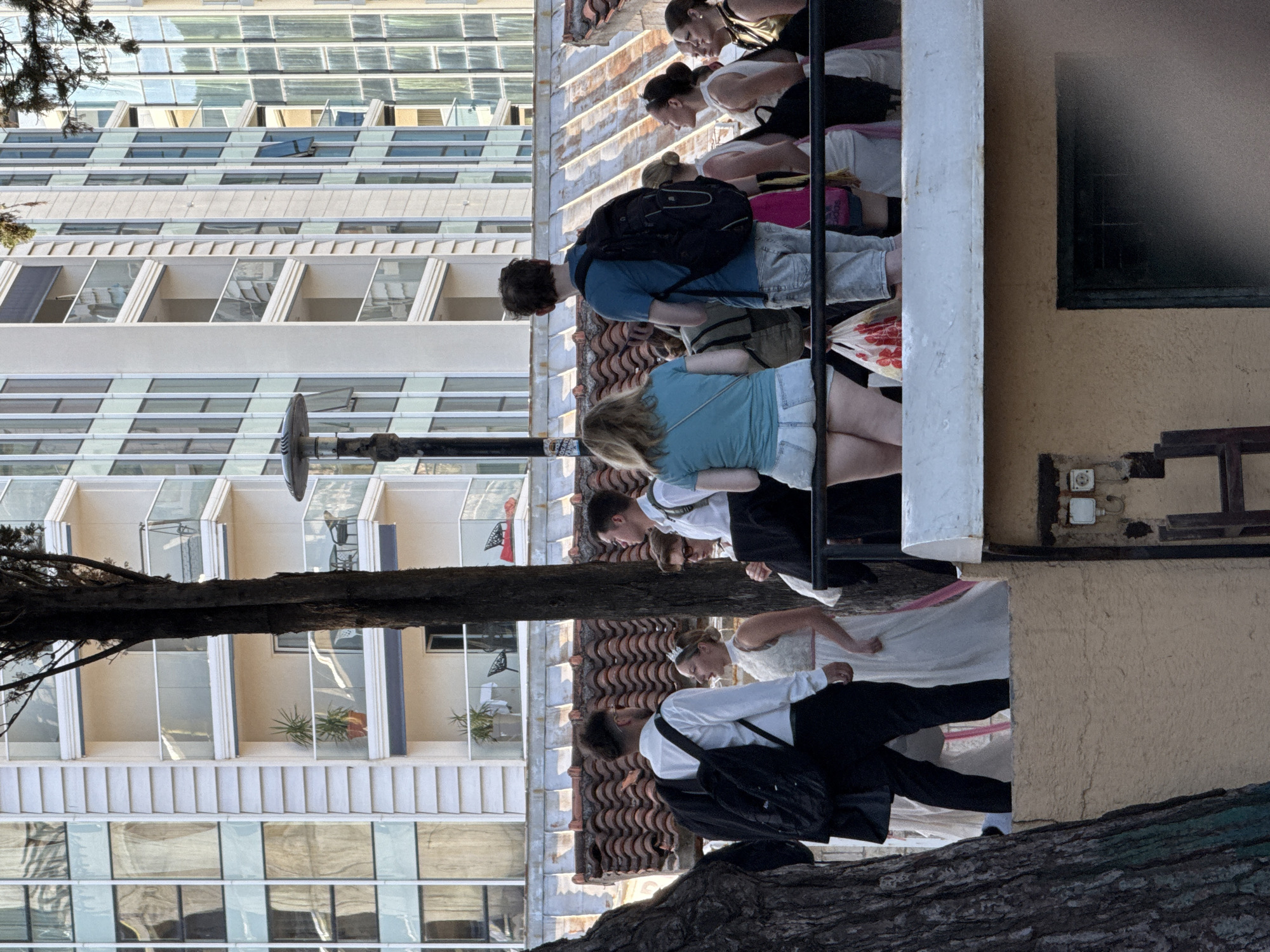 This image captures a group of approximately twelve individuals gathered on an elevated outdoor platform or terrace in the city of Budva, Montenegro. The scene is set against a backdrop of modern high-rise buildings.
In the immediate foreground, a thick tree trunk occupies the lower-left section of the frame. Below the platform, a textured light brown wall is visible, with a small, light-colored utility box featuring two electrical outlets mounted on it in the lower-right. A black metal railing extends horizontally across the platform, and a dark lamppost with a broad top stands behind it on the left.
The individuals on the platform, predominantly young adults, are arranged in two main clusters. On the left, a man is seen from behind, wearing a white long-sleeved shirt, black trousers, and a black backpack, with a black jacket draped over his left arm. To his right, a woman in a white, sleeveless formal dress with a pink detail at the waist and a small, light-colored headpiece is looking towards him with her mouth open. Another man in a white shirt and black backpack is partially visible behind the first man. Further right, a woman in a light blue t-shirt and light denim shorts is seated on the metal railing, facing away from the camera. A white plastic bag with red floral patterns hangs near her on the railing. Adjacent to her, a man in a blue t-shirt, light jeans, and a black backpack stands facing away. To their right, several other individuals, mostly women, are observed, some wearing white or gold-toned tops, with their gazes directed towards the right side of the frame.
Behind the group, a rooftop with distinctive terracotta-colored, wavy tiles is visible. The background is composed of multiple modern, multi-story buildings characterized by numerous large, reflective glass windows and light-colored facades. Several balconies with glass railings are evident on these structures, some containing potted plants. A second, thinner dark tree trunk rises vertically in the mid-left background, partially obscuring one of the background buildings. The scene is illuminated by bright, clear sunlight, indicating either late morning or late afternoon conditions.