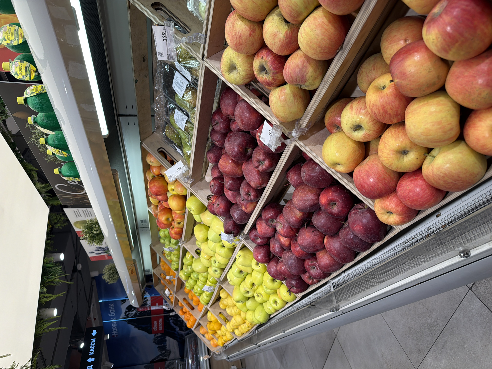 The image depicts a produce section in a store, likely a grocery store, located in Khimki, Russia, as evidenced by signage in Russian. The display shelves are organized with a variety of fruits. Apples in shades of red, yellow and mixed colors are arranged in separate wooden crates at the top. Pears in plastic packaging are on the upper left shelf. Moving downward, there are oranges and yellow apples. The lighting is bright and even, illuminating the merchandise. The overall scene is a typical grocery store setup with fruits neatly displayed. The floor is tiled, and part of the ceiling is visible, showing additional store elements such as signs and greenery.
