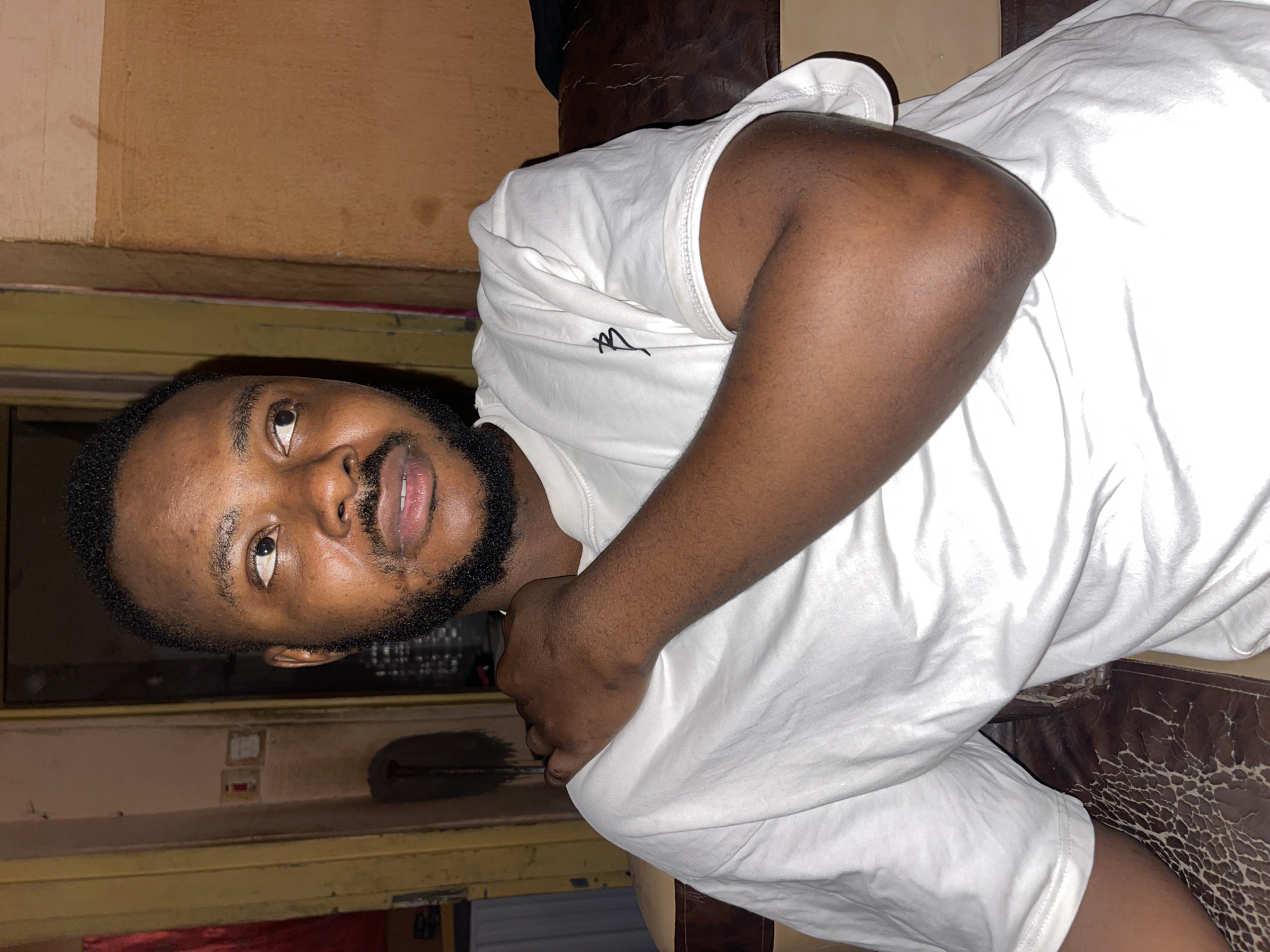 A man is lying on a patterned surface, likely a damaged leather sofa, in an interior setting. He is wearing a white t-shirt. The background appears to be a wall, suggesting a room indoors. The location is unknown.