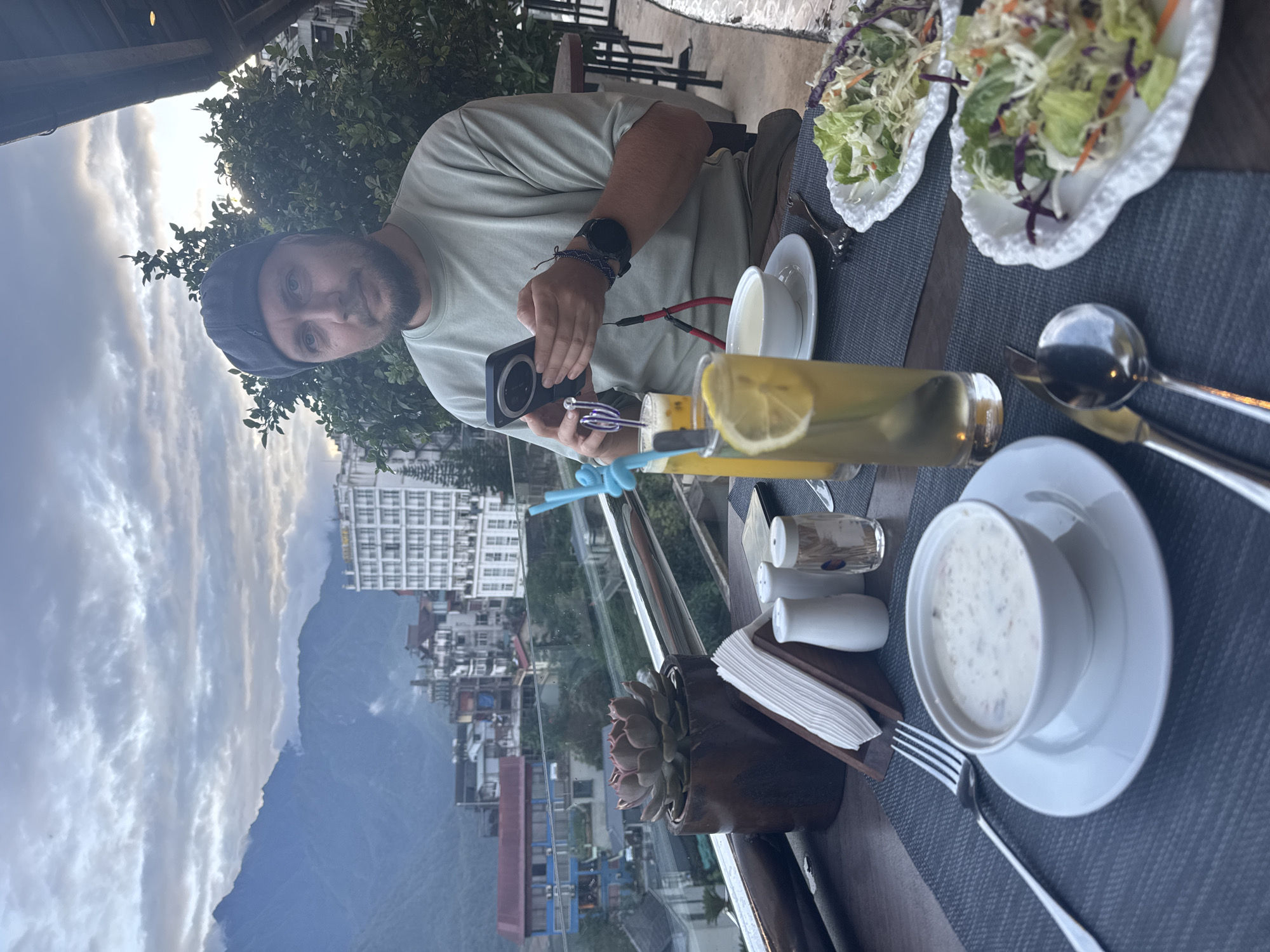 A man wearing a gray cap and light green shirt is at a table in what appears to be a restaurant or patio setting. He is holding a smartphone in his hands, seemingly taking a photo. On the table are two salads, a drink with lemon slices, a bowl of soup, and a small plant in a wooden container. Salt and pepper shakers and napkins are also visible. A landscape is visible in the background. The location is Sa Pa Ward, Vietnam, based on the architectural style of the buildings and mountainous terrain.