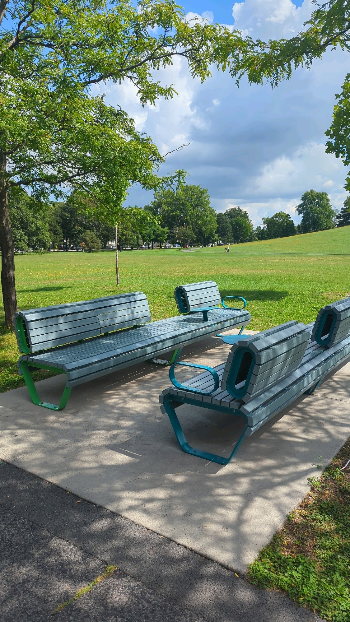 The image depicts a park scene with two modern, teal-colored benches with green accents positioned on a concrete paved area. The benches are facing a large, grassy field that extends into the distance, with a gentle slope on the right side of the field. Mature trees with lush green foliage are scattered throughout the park, with a prominent tree on the left side of the frame partially obscuring the sky. The sky is partly cloudy with dramatic, dark clouds interspersed with patches of blue. In the middle distance, two figures are walking across the grassy field. The ground in the foreground is a mix of paved surfaces and grass, with shadows cast by the trees. The scene suggests a public park in an urban setting, likely Montreal, Canada, given the presence of typical urban park features and landscaping.