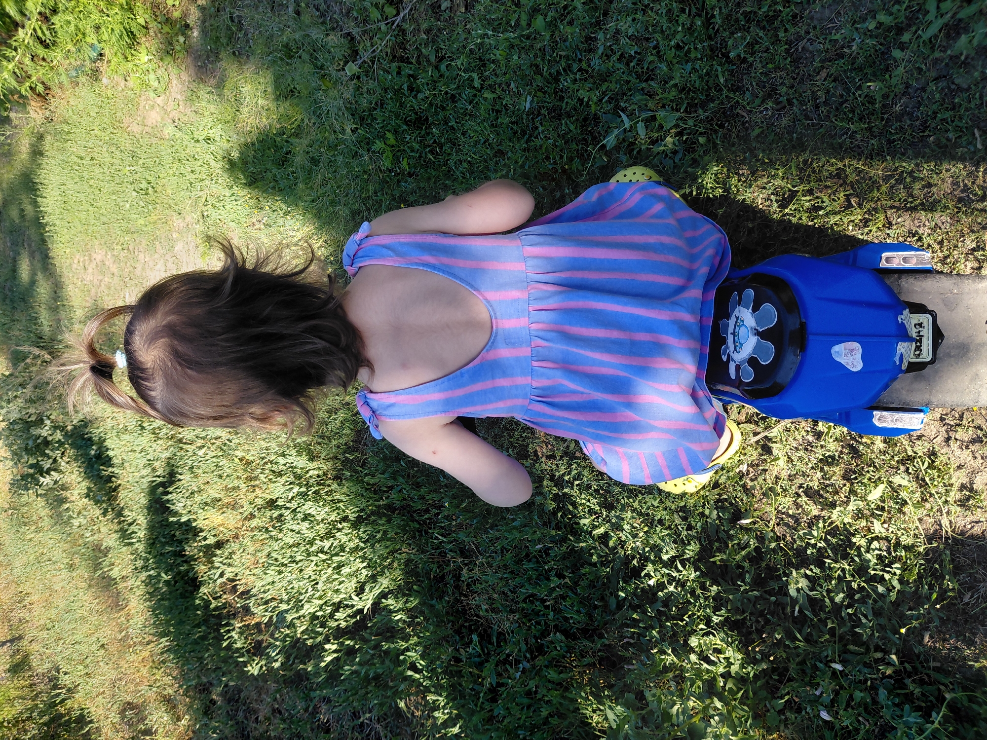 The image shows a young child lying on grass next to a blue toy vehicle. The child is wearing a blue and pink striped dress and yellow shoes. Their hair is pulled back in a ponytail. The toy vehicle appears to be a ride-on toy with wheels and a steering wheel. The scene is set outdoors, with green grass and some shadows suggesting it is daytime. The child is positioned on their stomach, facing away from the camera, and their arm is resting on the grass. The toy vehicle is in front of the child. The ground is covered in grass and some patchy dirt. There are no other people visible in the image. The lighting suggests it is a sunny day.