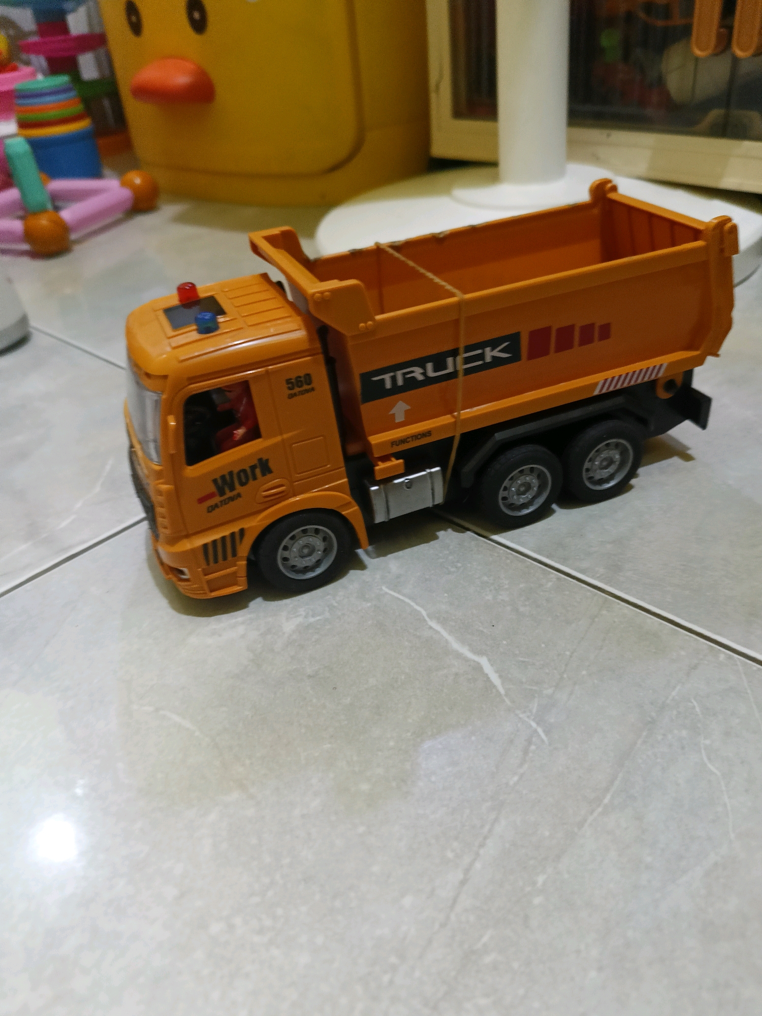 An orange toy dump truck is positioned on a tiled floor. The truck has the word "TRUCK" printed on its side, along with the number "560" and "Work". It features red and blue lights on the roof of the cab. In the background, blurred toys and a portion of a white fan are visible. The scene is indoors, likely in a residential setting. No people are visible in the image. The location is not specified as Tangerang, Indonesia.